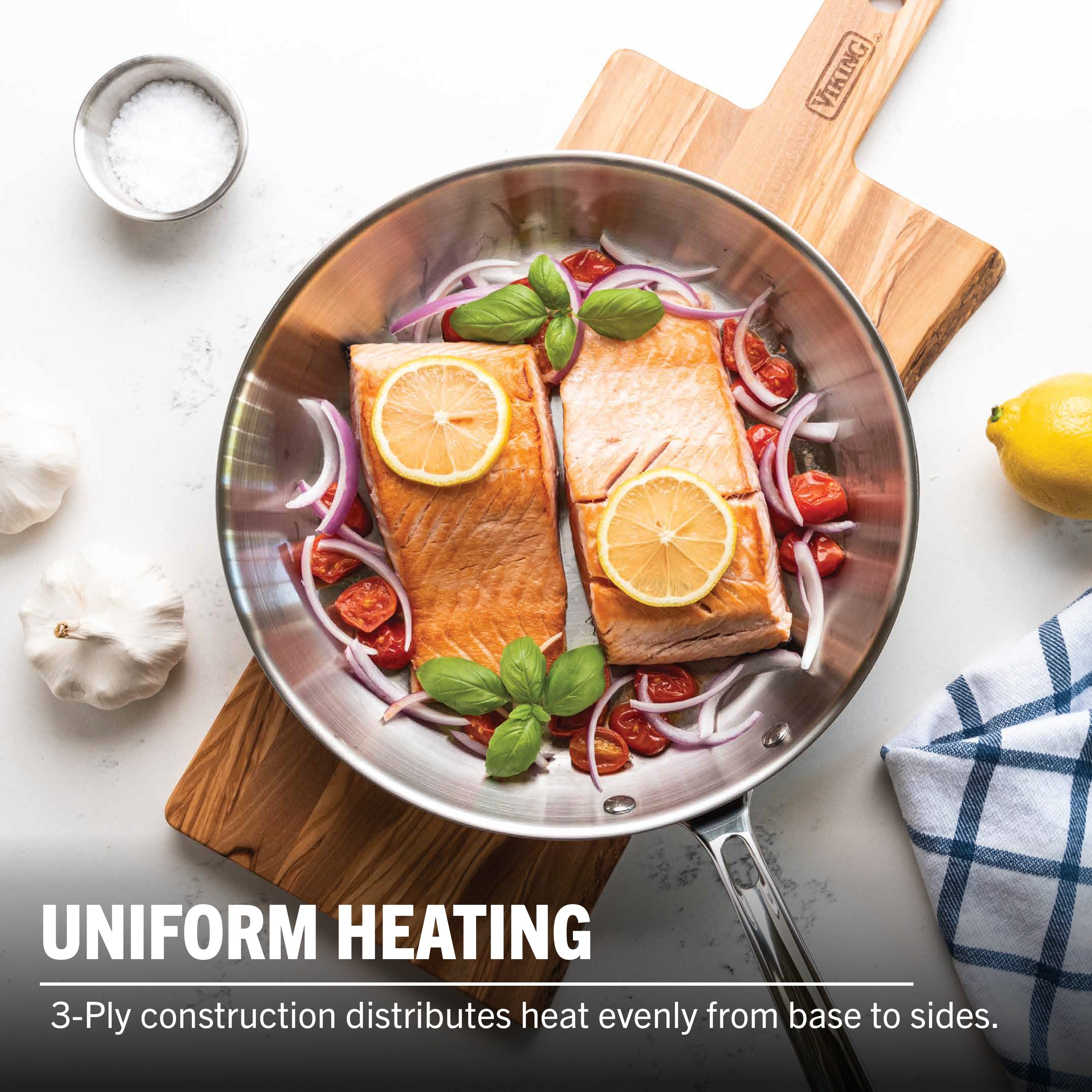 The Viking Artisan 3-Ply Stainless Steel 10-Inch Fry Pan by Viking Culinary Products holds two salmon fillets with lemon, red onions, cherry tomatoes, and basil. Garlic, lemon, salt in a metal bowl, and a blue-checkered cloth surround it. Uniform Heating. Induction Compatible.