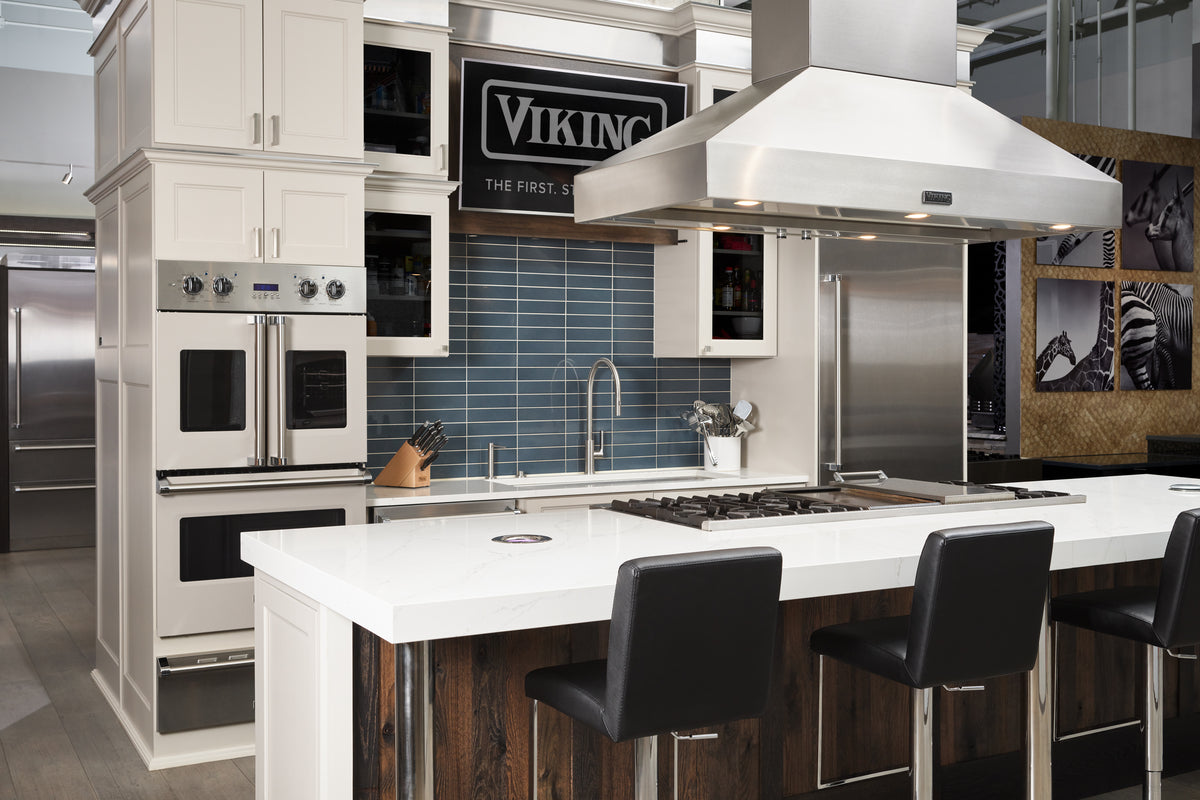 Modern kitchen with white cabinets, blue tile backsplash, stainless steel appliances, a large island with a stove and range hood, and three black barstools.