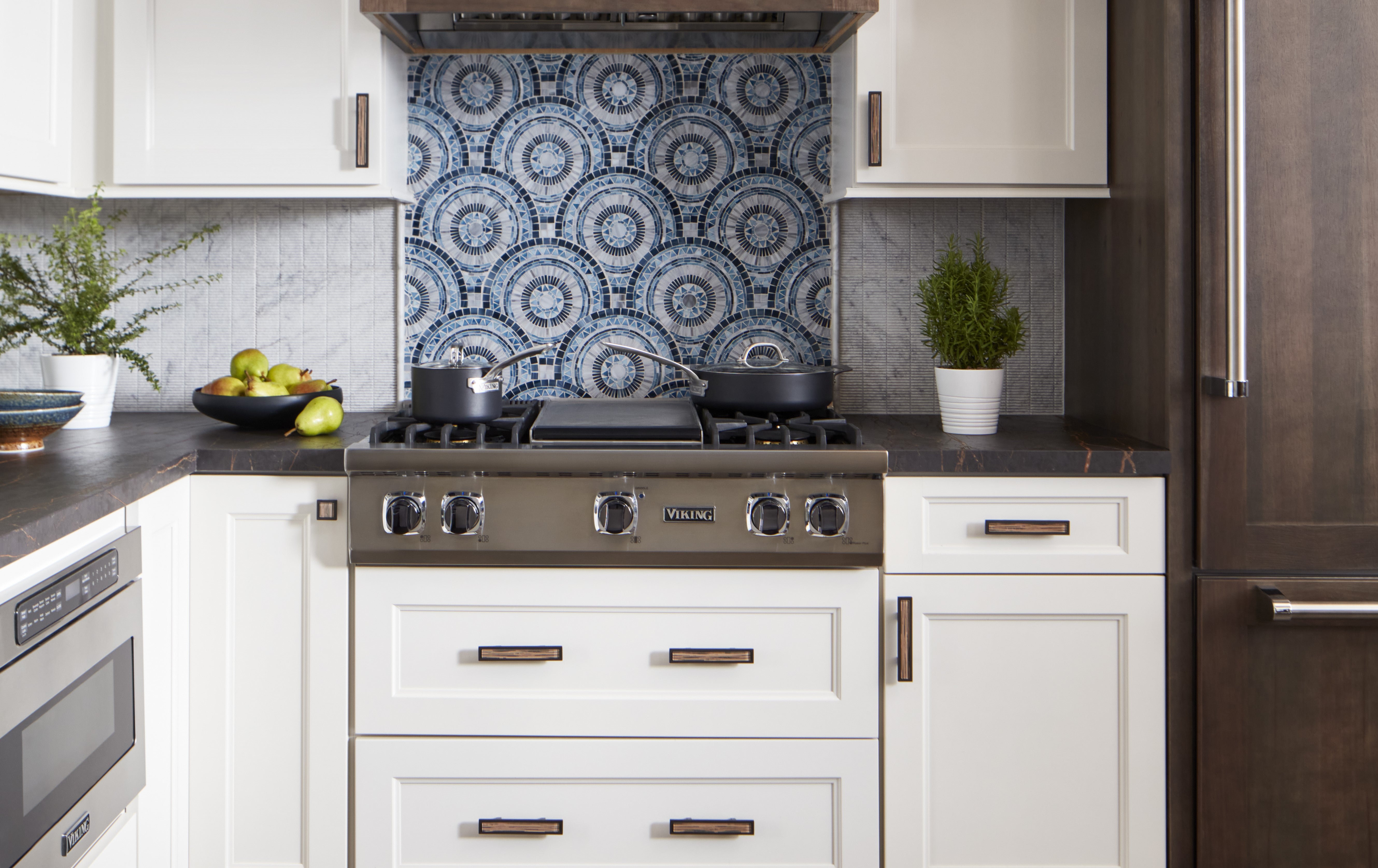 A modern kitchen with a stainless steel stove, white cabinets, a blue patterned tile backsplash, two potted plants, a bowl of green apples, and dark countertops.