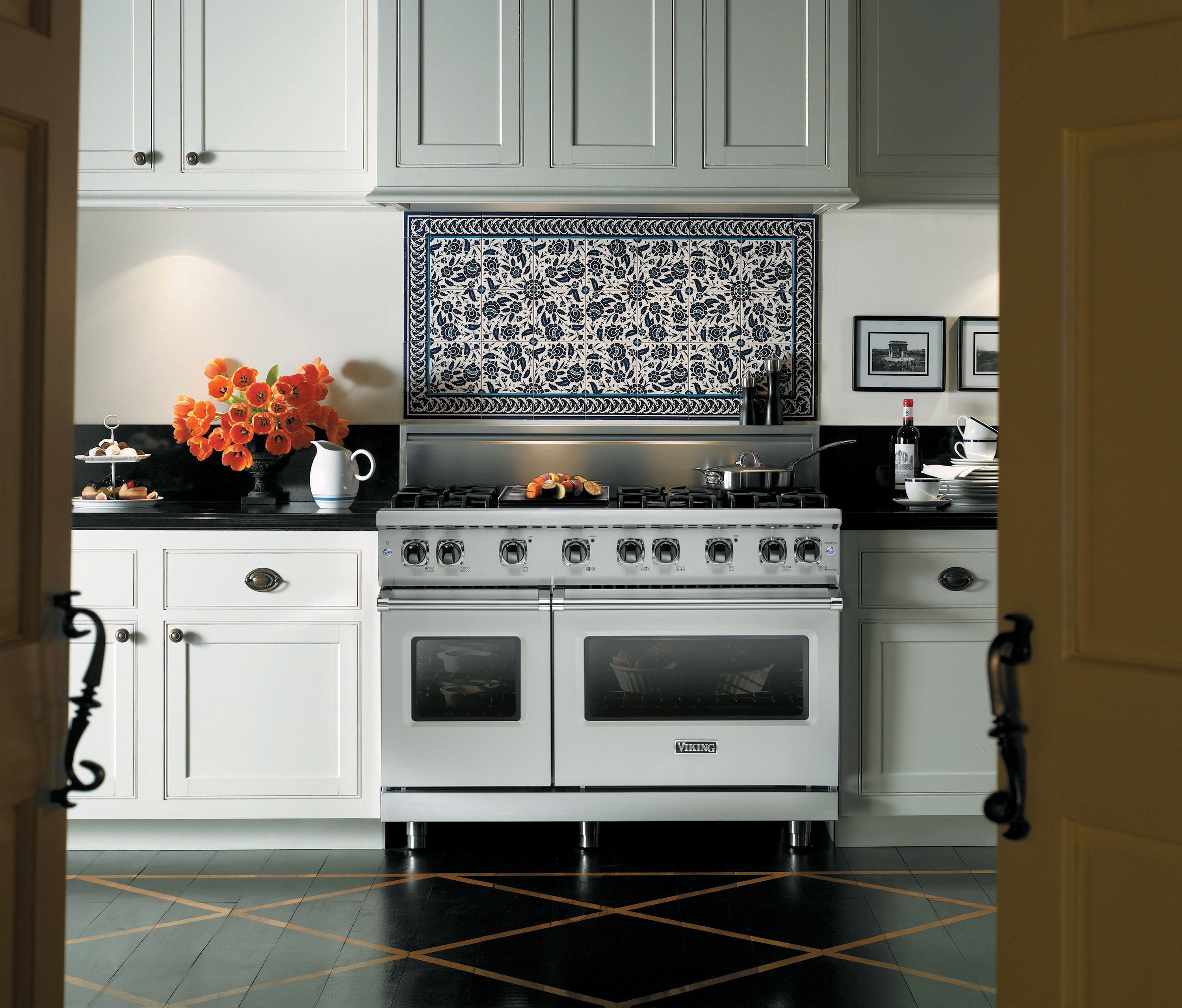 A modern kitchen with white cabinets, black countertops, a stainless steel oven, a mosaic tile backsplash, orange flowers in a vase, and plates of food on the counter.