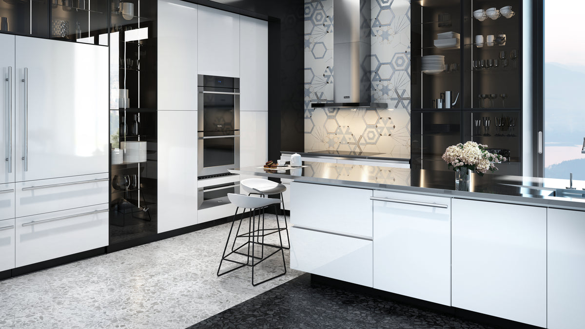 Modern kitchen with glossy white cabinets, built-in ovens, a large island with a sink, bar stools, and glass shelving displaying dishes. Flowers in a vase are on the island, and a window shows a scenic view.