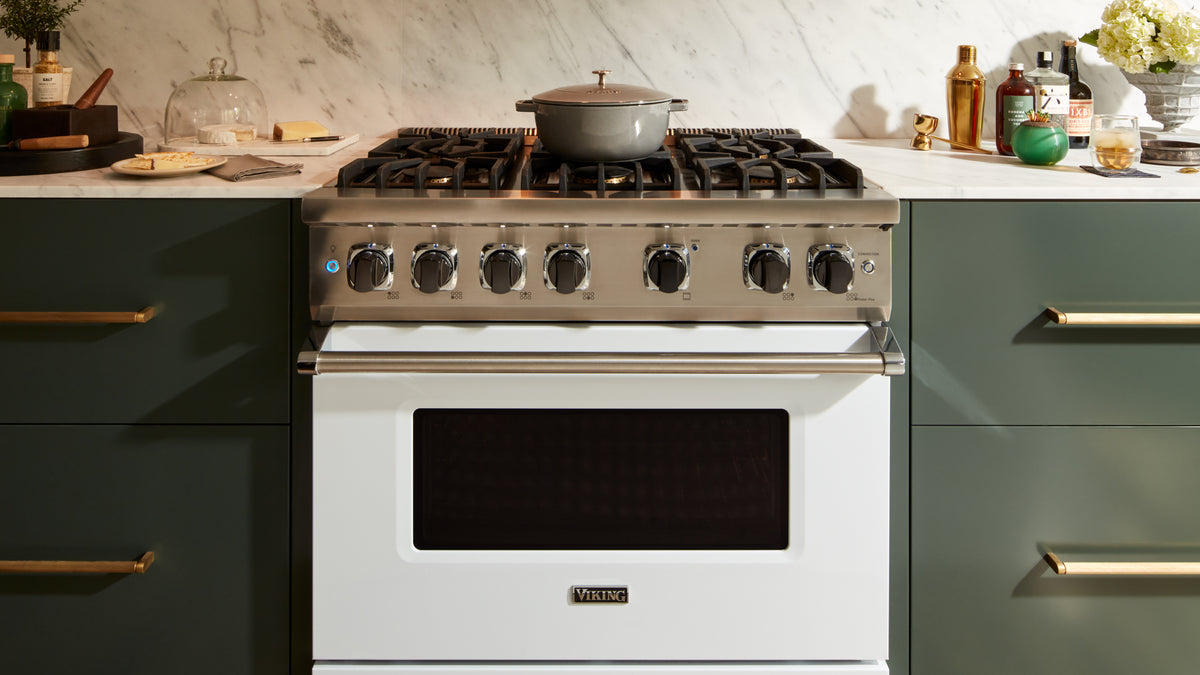 A stainless steel gas stove with six burners and a white oven, set between green cabinets with a marble backsplash. A pot is on the center burner, and various kitchen items are on the countertop.