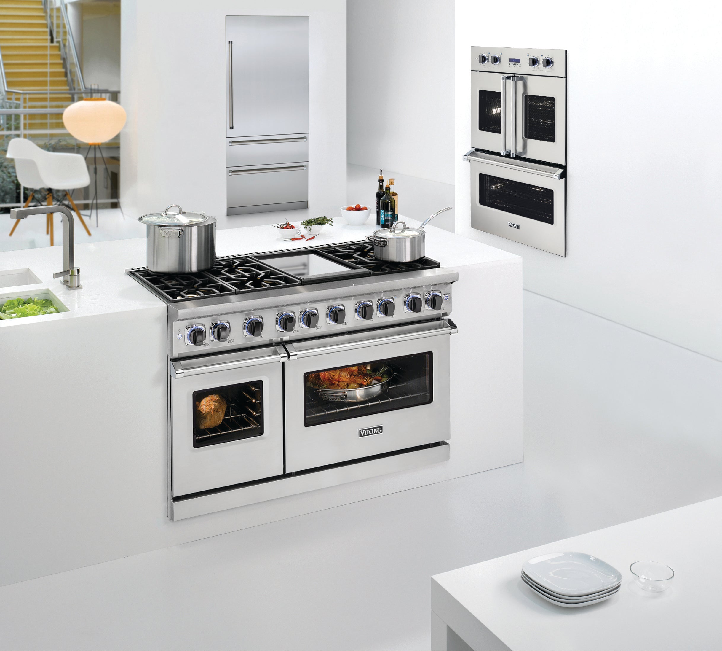 Modern, sleek white kitchen with a stainless steel stove featuring double ovens, two pots on the stovetop, a built-in wall oven, and minimalistic decor. Plates are set on a white counter in the foreground.