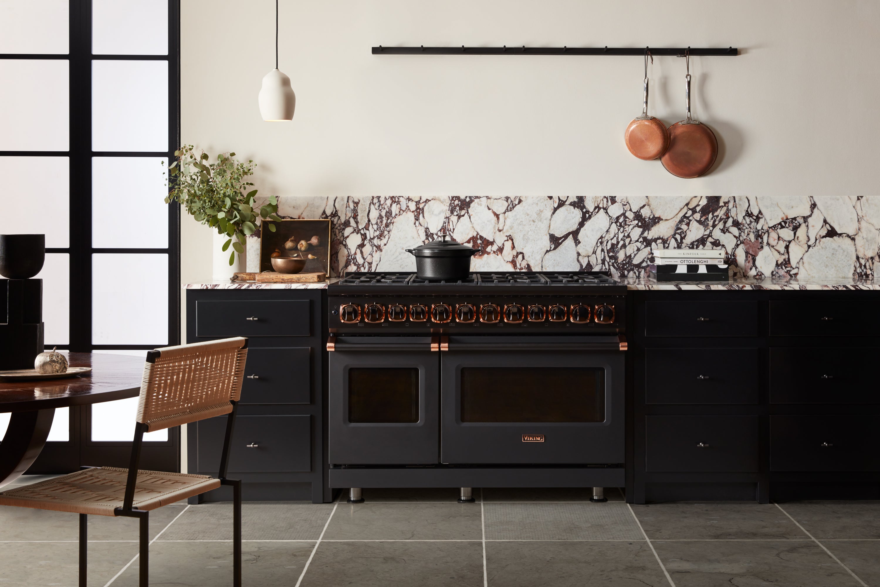 A modern kitchen with black cabinets, a large black stove, marble backsplash, hanging copper pans, a plant, and a small dining table with a wicker chair. The space is sleek and minimalist with neutral tones.