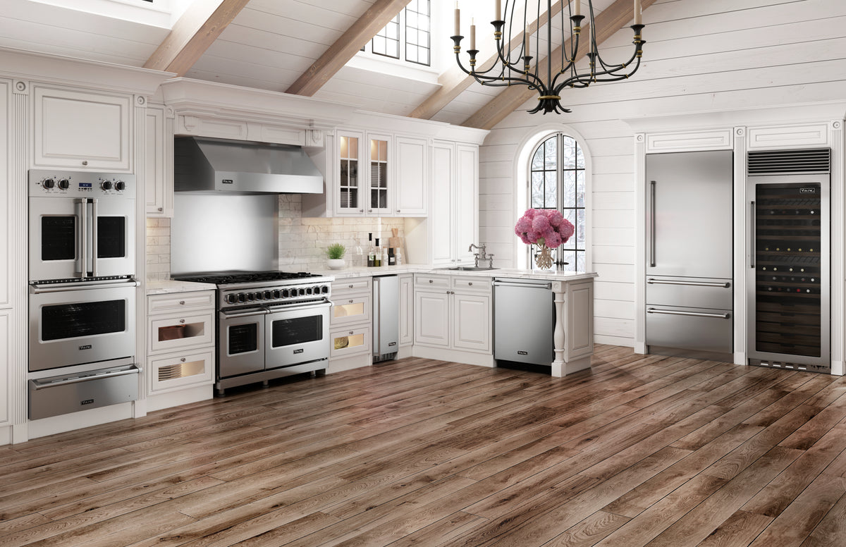 Spacious, elegant kitchen with white cabinetry, stainless steel appliances, wood floors, a large range, double ovens, wine fridge, farmhouse sink, and a black chandelier under a high ceiling with skylights. Pink flowers decorate the counter.