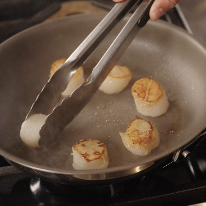 A person uses tongs to sear scallops in a Viking PureGlide Pro 3-Ply 8 Inch Fry Pan by Viking Culinary Products on a stovetop; the scallops are browning as steam rises.