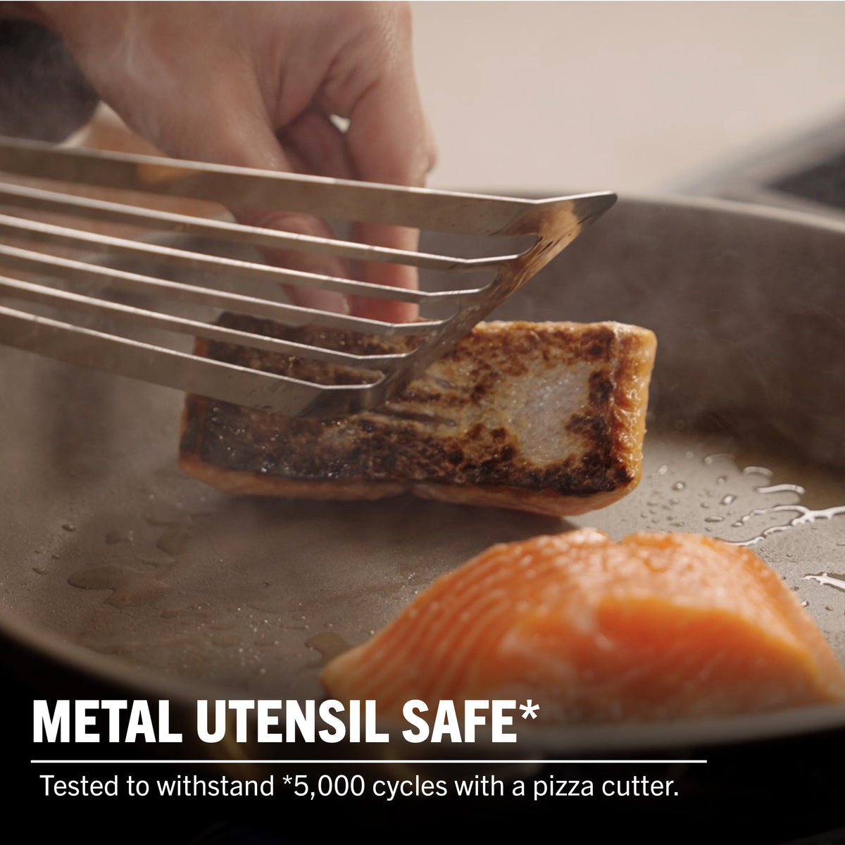 A hand uses a metal spatula to lift cooked fish from the Viking PureGlide Pro 3-Ply Fry Pan, with raw salmon beside it. Text: METAL UTENSIL SAFE*—withstands 5,000 cycles with a pizza cutter. Viking Culinary Products.