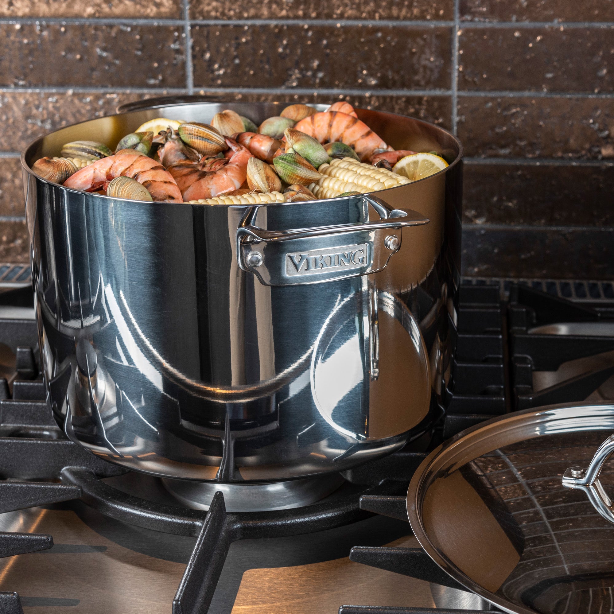 The Viking Culinary Products 3-Ply Stainless Steel 12-Quart Stock Pot with Metal Lid sits on a stovetop, filled with shrimp, clams, and pasta. Its lid rests to the side against a backdrop of brown tiled wall.