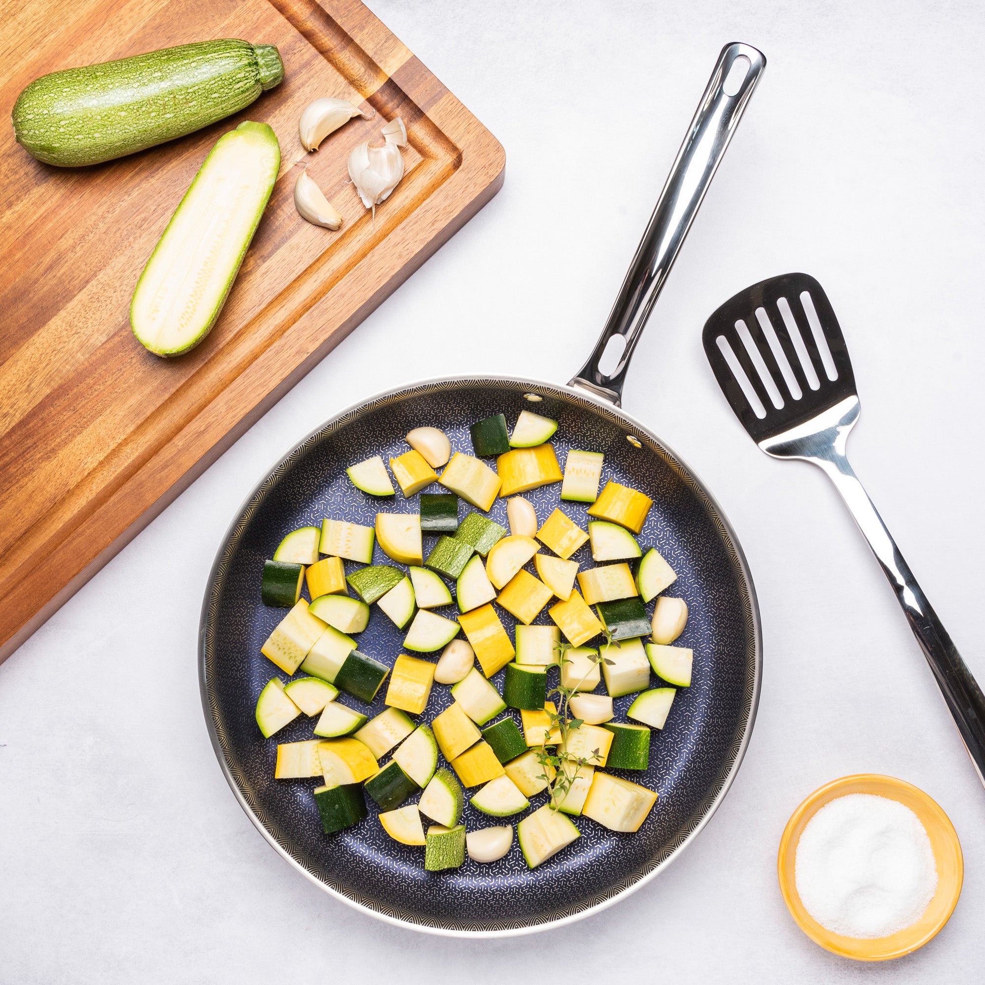 A Viking Culinary Products 3-Ply Hybrid Plus 12-inch Nonstick Fry Pan holds chopped green and yellow zucchini on a white surface, with a spatula, salt bowl, garlic cloves, knife, and cutting board with whole and halved zucchini nearby.