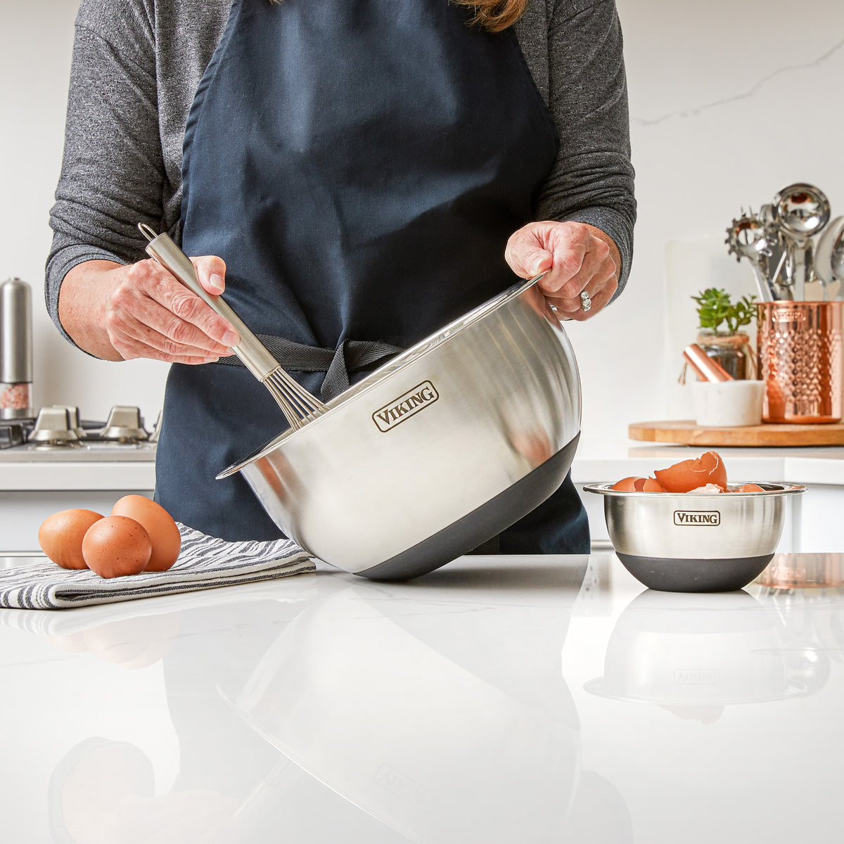 A person in a dark apron whisks ingredients in a Viking 10-Piece Stainless Steel Mixing Bowl with a black silicone base by Viking Culinary Products. Nearby are eggs, a smaller bowl, and kitchen utensils on the counter.