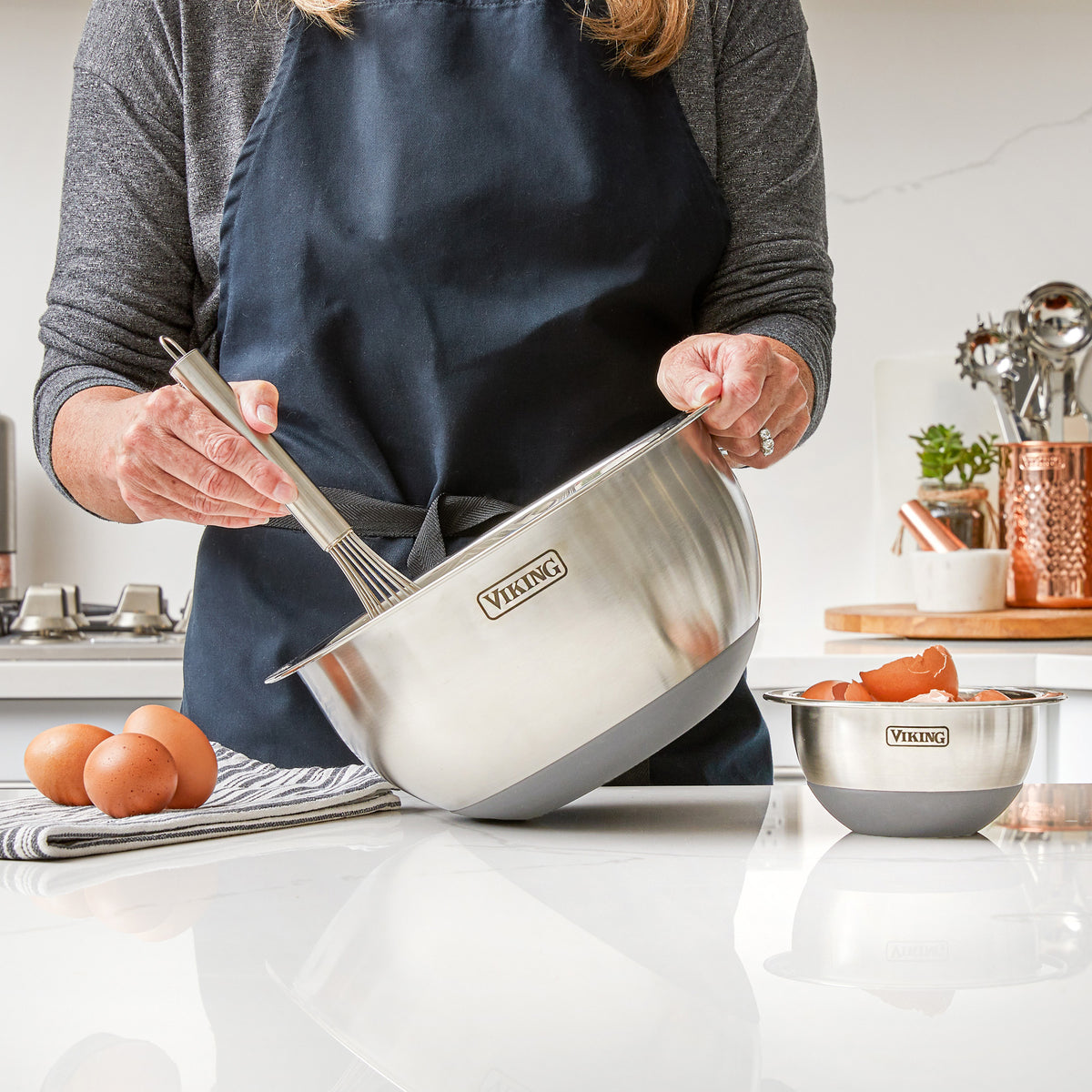 A person in a dark apron is whisking ingredients in a large bowl from the Viking 10-Piece Stainless Steel Mixing Bowl Set with Lids by Viking Culinary Products, with eggs, a smaller bowl, utensils, and compact storage containers on the kitchen counter.