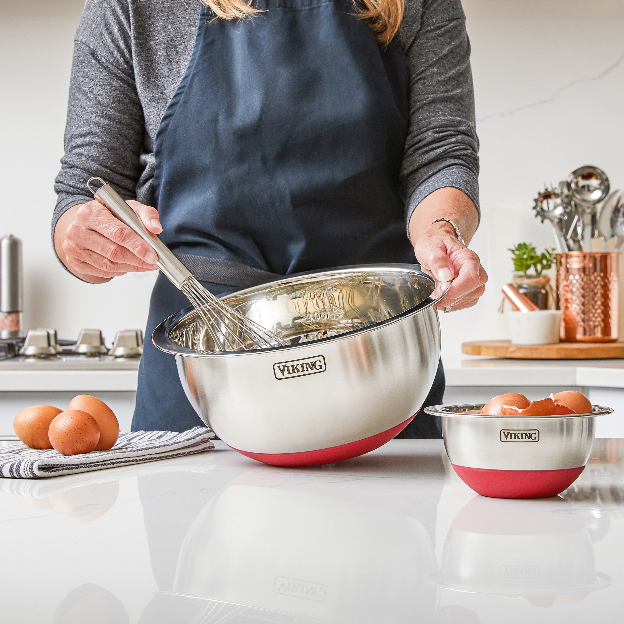 Wearing a navy apron, a person whisks eggs in a stainless steel mixing bowl with silicone base from the Viking 10-Piece Stainless Steel Mixing Bowl Set with Lids by Viking Culinary Products; another bowl and utensils are nearby.