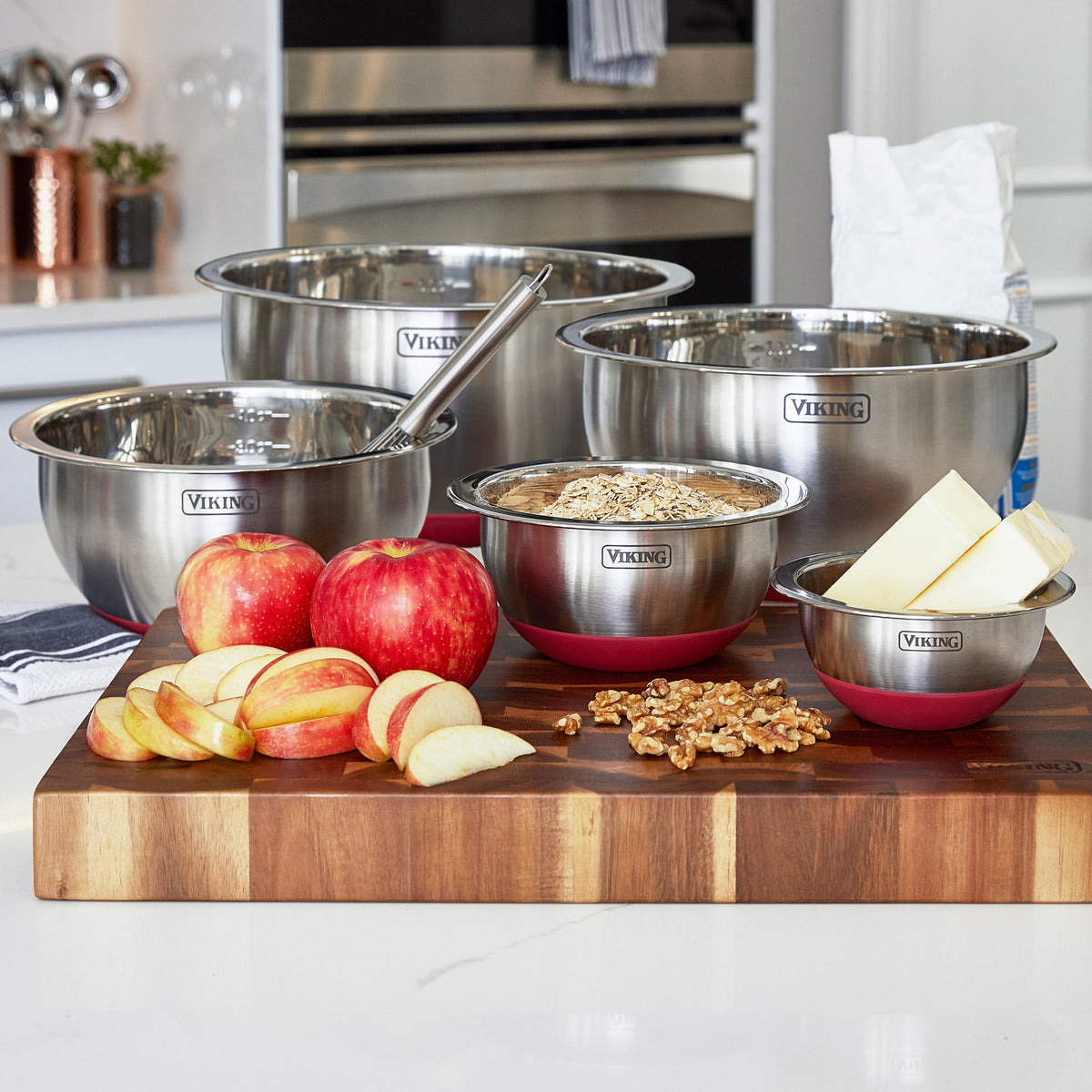 The Viking 10-Piece Stainless Steel Mixing Bowl Set with Lids by Viking Culinary Products is shown on a kitchen counter holding apples, walnuts, butter, and flour. A whisk rests in one bowl—ideal for all your baking and prep needs.