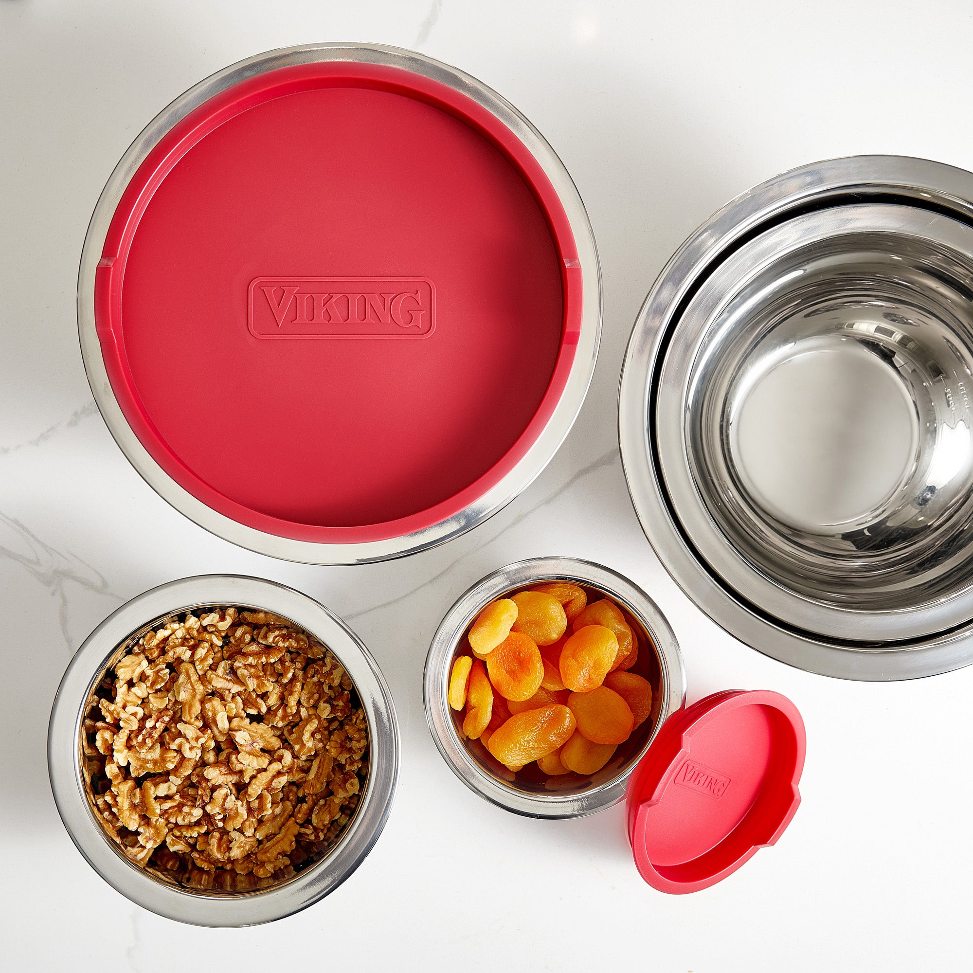 Three Viking Culinary stainless steel mixing bowls from the Viking 10-Piece Set, each with red silicone bases—one topped with a red lid, one filled with walnuts, one with dried apricots—are displayed on a white surface.