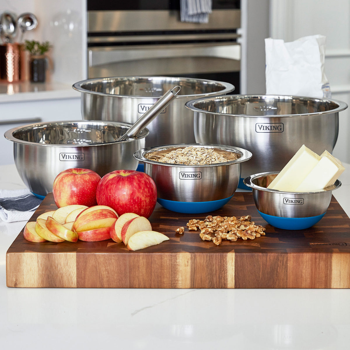 The Viking 10-Piece Stainless Steel Mixing Bowl Set with Lids by Viking Culinary Products sits on a kitchen counter, surrounded by apples, butter, walnuts, oats on a wooden cutting board, and a nearby bag of flour for easy storage.