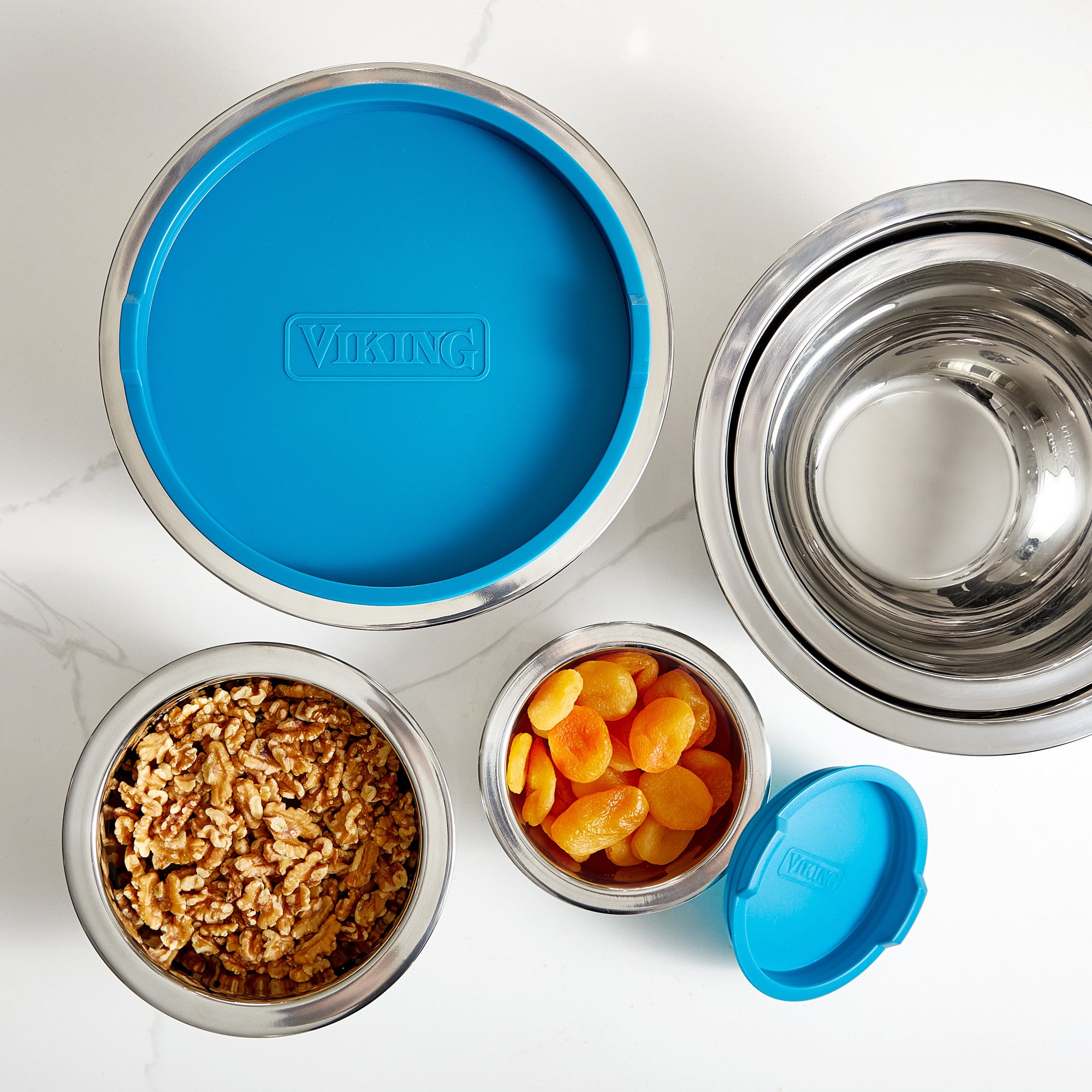 The Viking 10-Piece Stainless Steel Mixing Bowl Set with Teal Lids by Viking Culinary Products is shown on a white surface. Two bowls have teal airtight lids labeled VIKING, one holds walnuts, another apricots; the other two are empty.