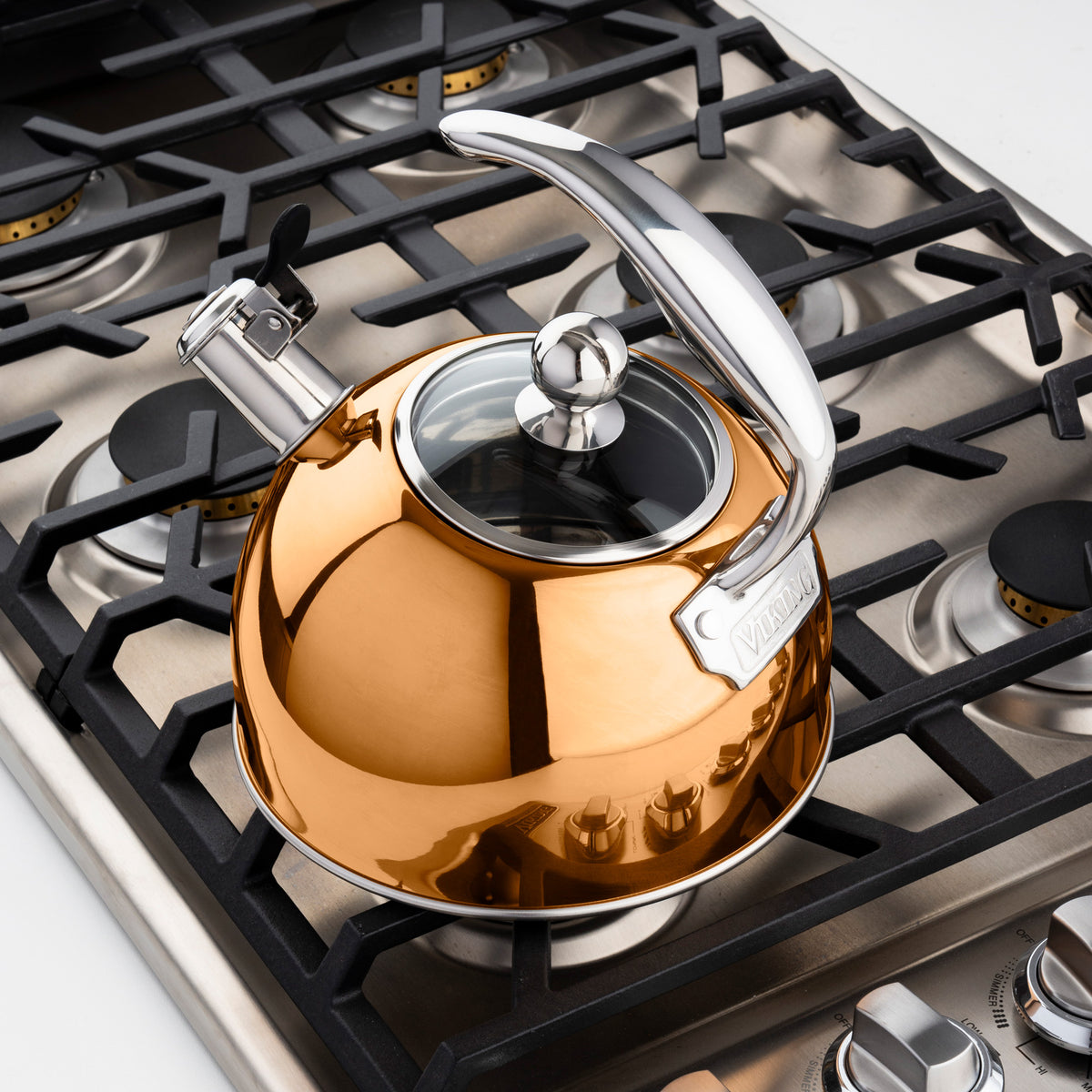 The Viking 2.6-Quart Rose Gold Stainless Steel Whistling Kettle by Viking Culinary Products, featuring a glass-topped lid and lever-whistle spout, sits on a stainless steel gas stove with black grates.