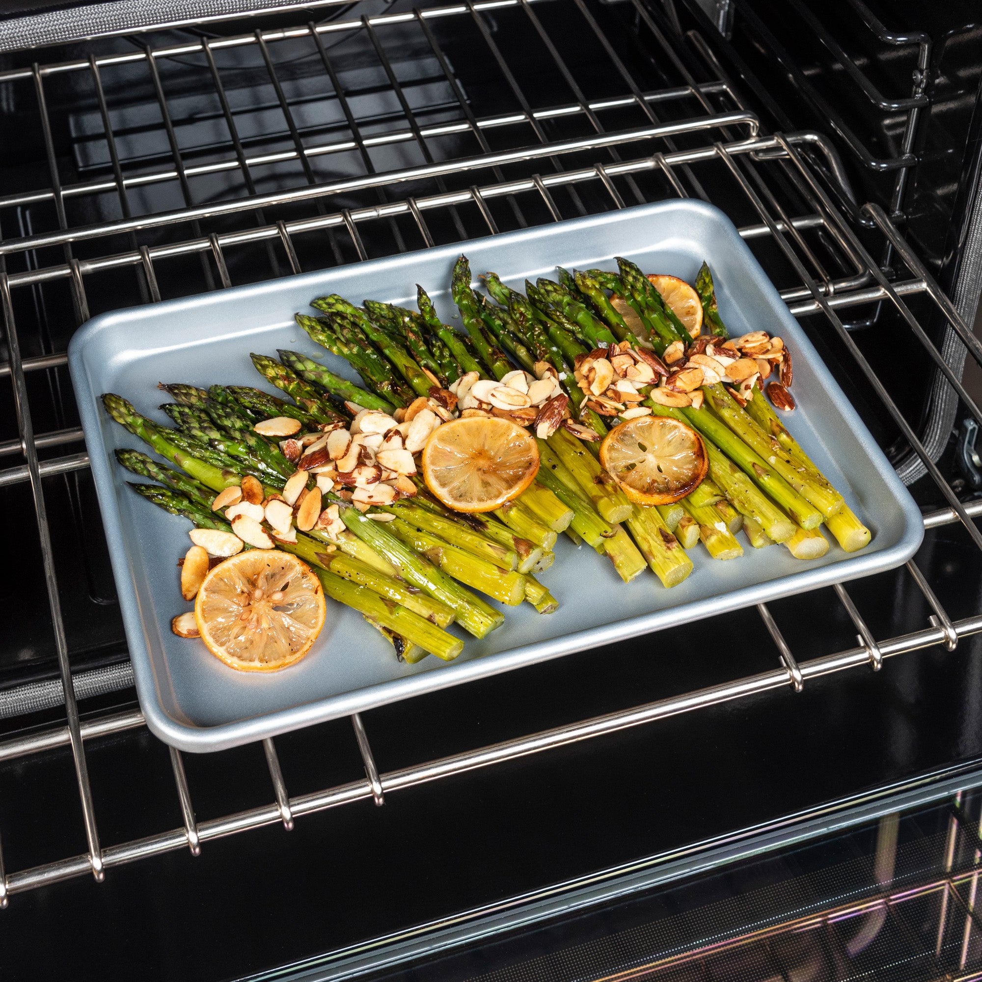 Roasted asparagus with lemon slices and almonds sits on a Viking 3-Piece Nonstick Aluminized Steel Baking Sheet from Viking Culinary Products, inside the oven.