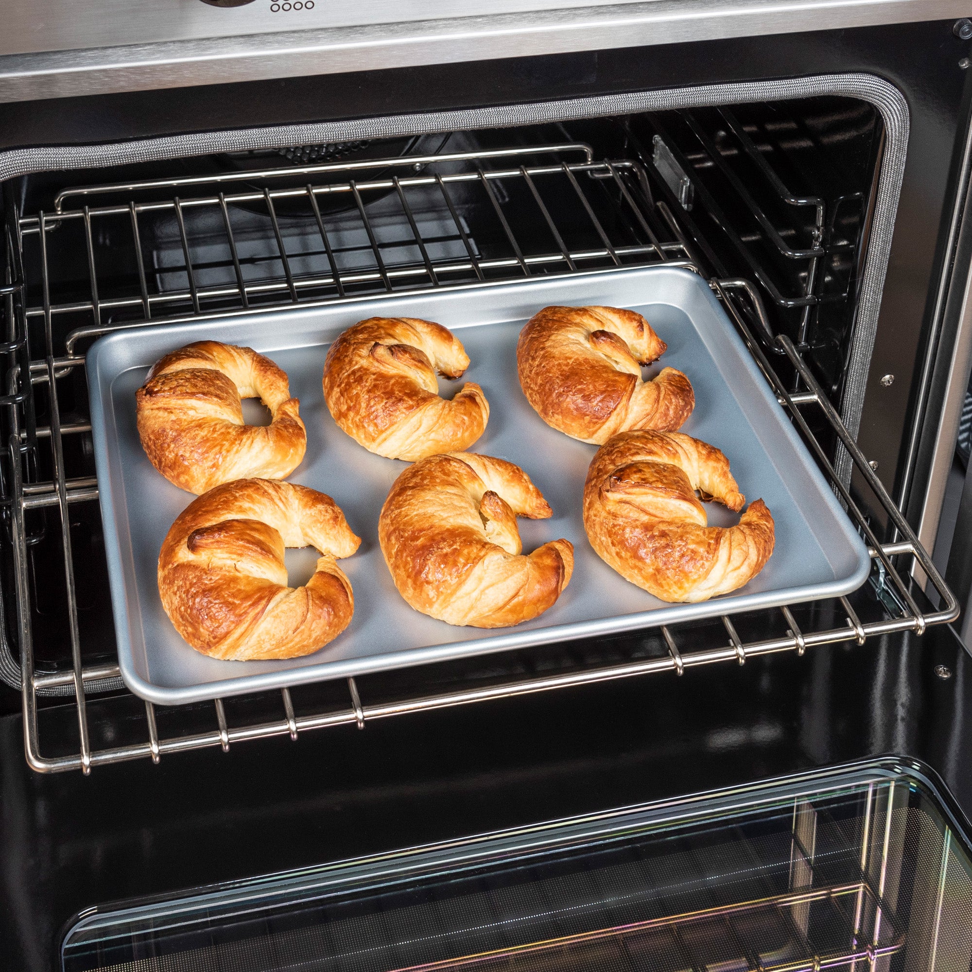 Six golden-brown croissants rest on a Viking 15-Inch Aluminized Nonstick Baking Sheet by Viking Culinary Products, placed inside an open oven on a metal rack.