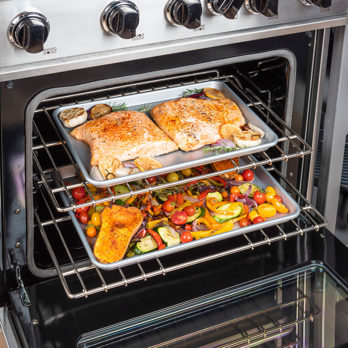 Two trays in the oven: the top holds seasoned salmon with veggies, the bottom has a colorful veggie mix—all baked evenly on Viking Culinary Products’ 2-Piece Nonstick Aluminized Steel Baking Sheet Set, known for excellent heat conductivity.