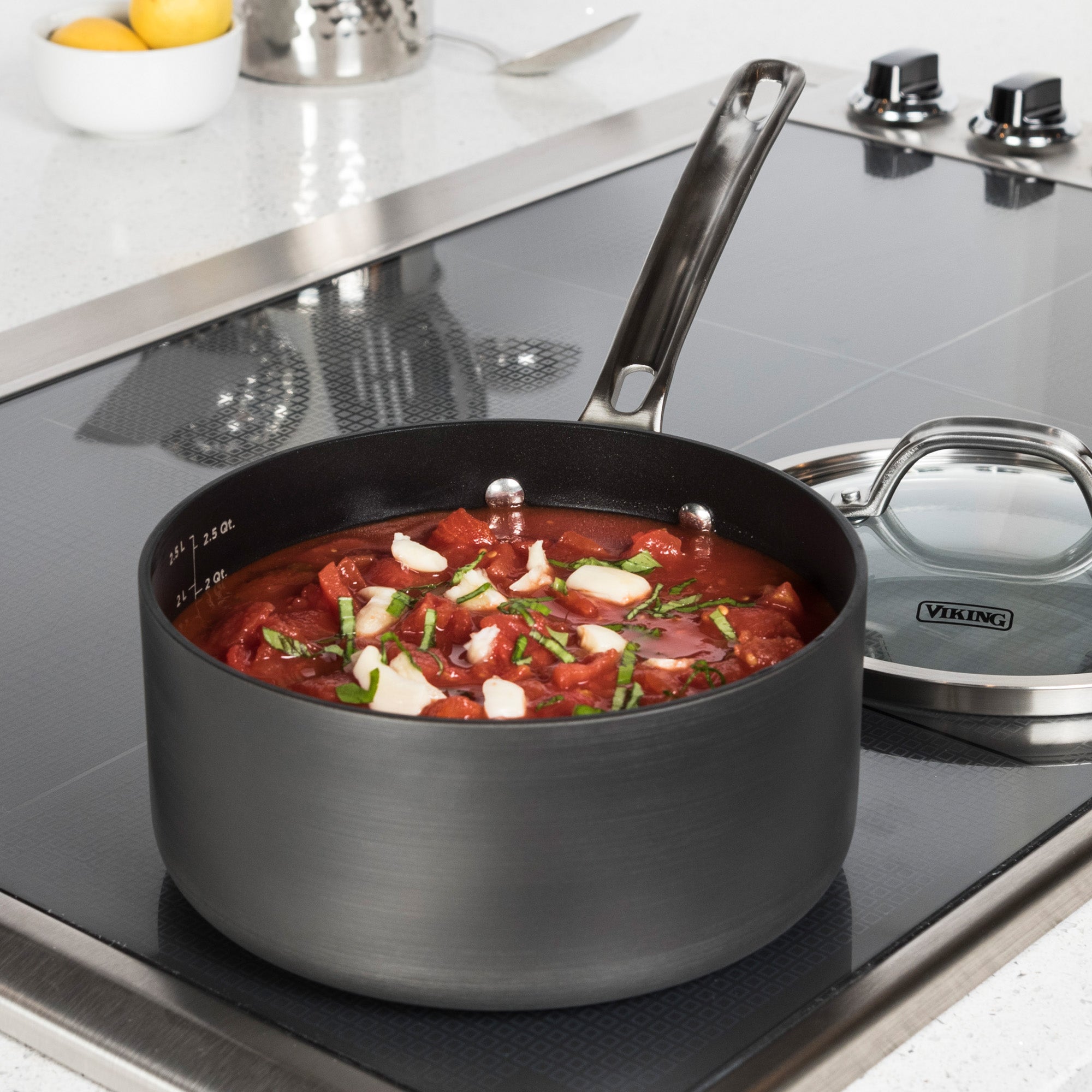 The Viking Hard Anodized Nonstick 3-Quart Sauce Pan by Viking Culinary Products, filled with tomato sauce, garlic, and herbs, sits on a modern stovetop. Its glass lid is nearby, with lemons and kitchen items in the background.