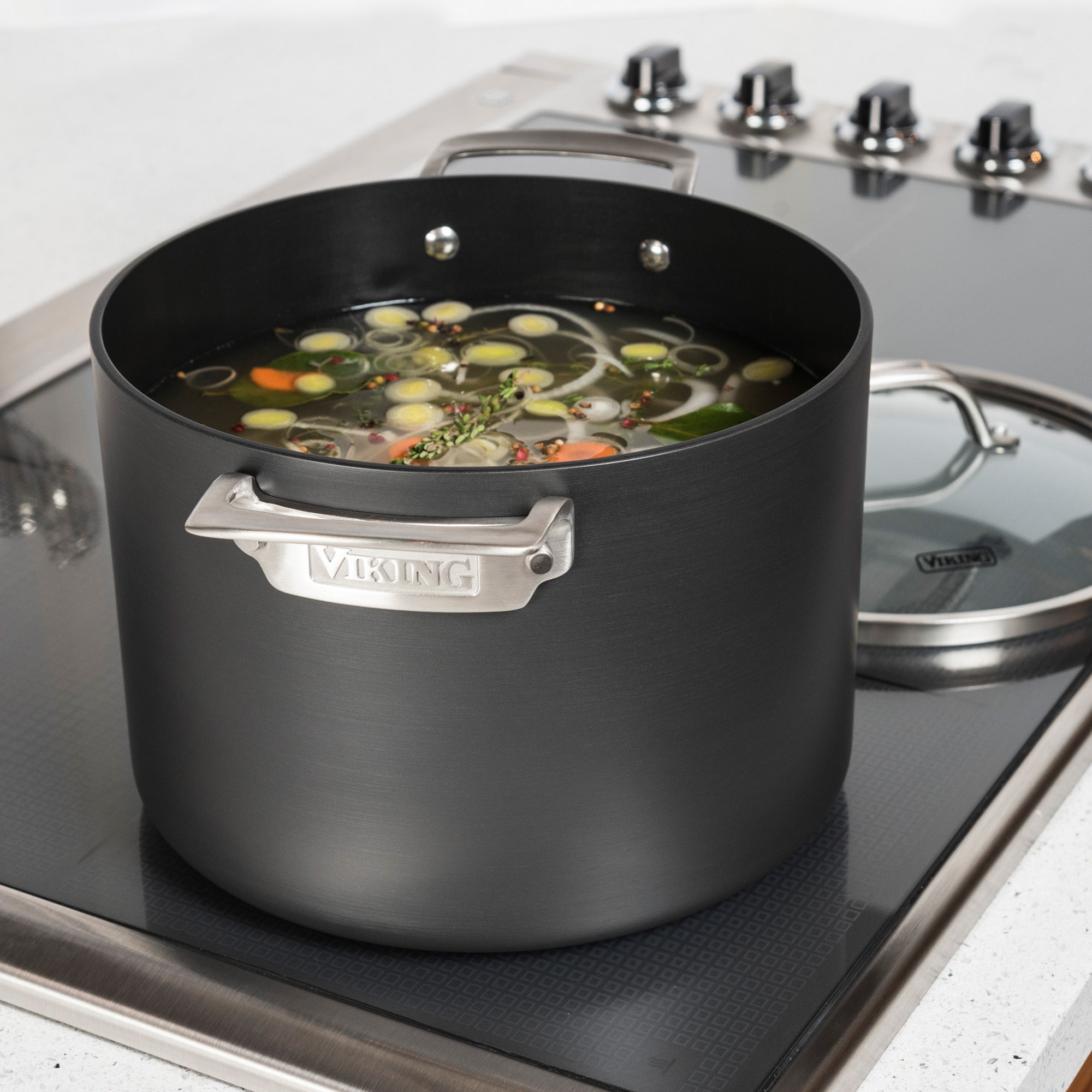 A Viking Hard Anodized Nonstick 8-Quart Stock Pot by Viking Culinary Products sits on the stove, filled with vegetable soup featuring sliced carrots, celery, leeks, and herbs in broth. The included glass lid rests nearby.