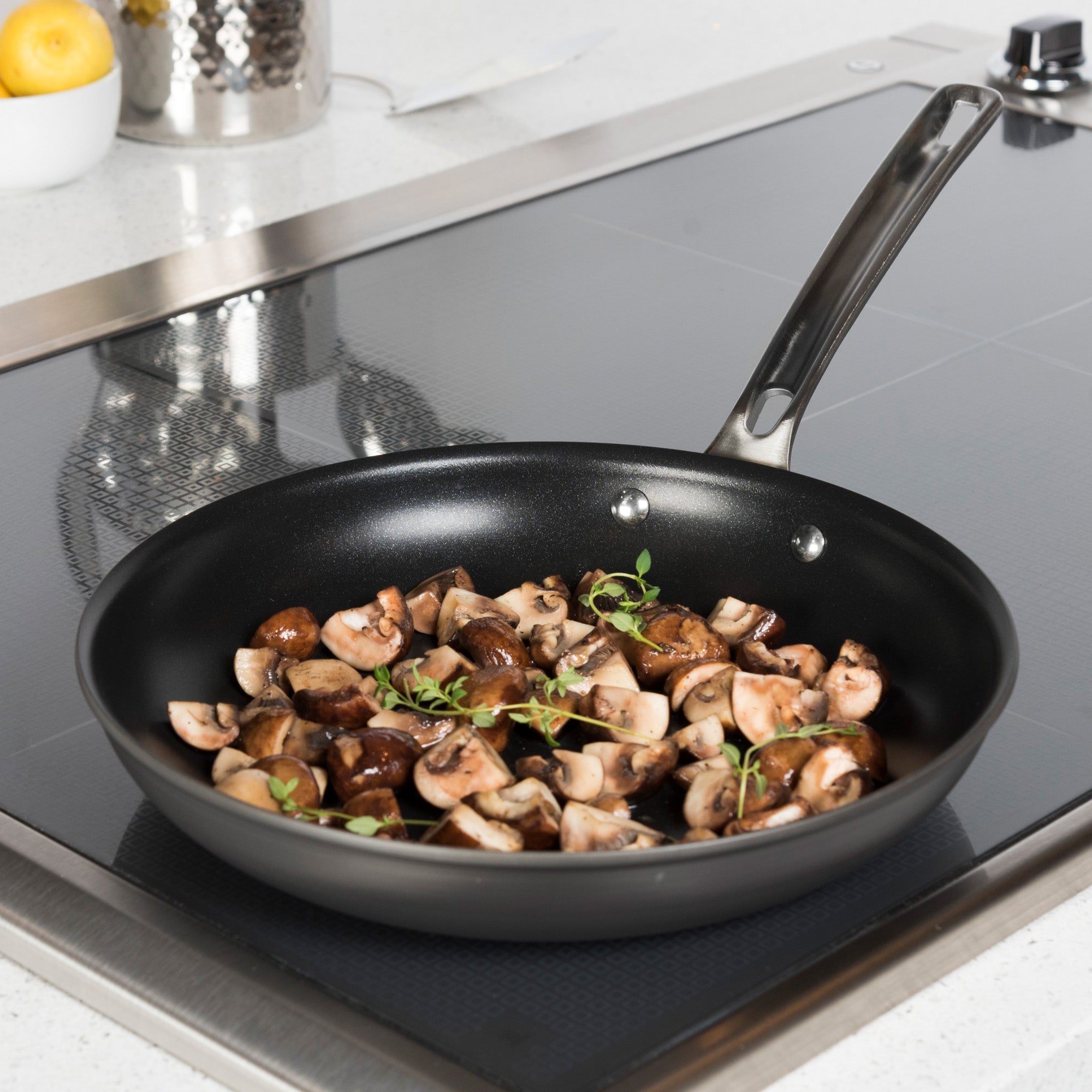 A Viking Culinary Products Hard Anodized Nonstick frying pan from the 10-piece set is shown on a stovetop in a modern kitchen, cooking chopped mushrooms and fresh herbs under glass lids.