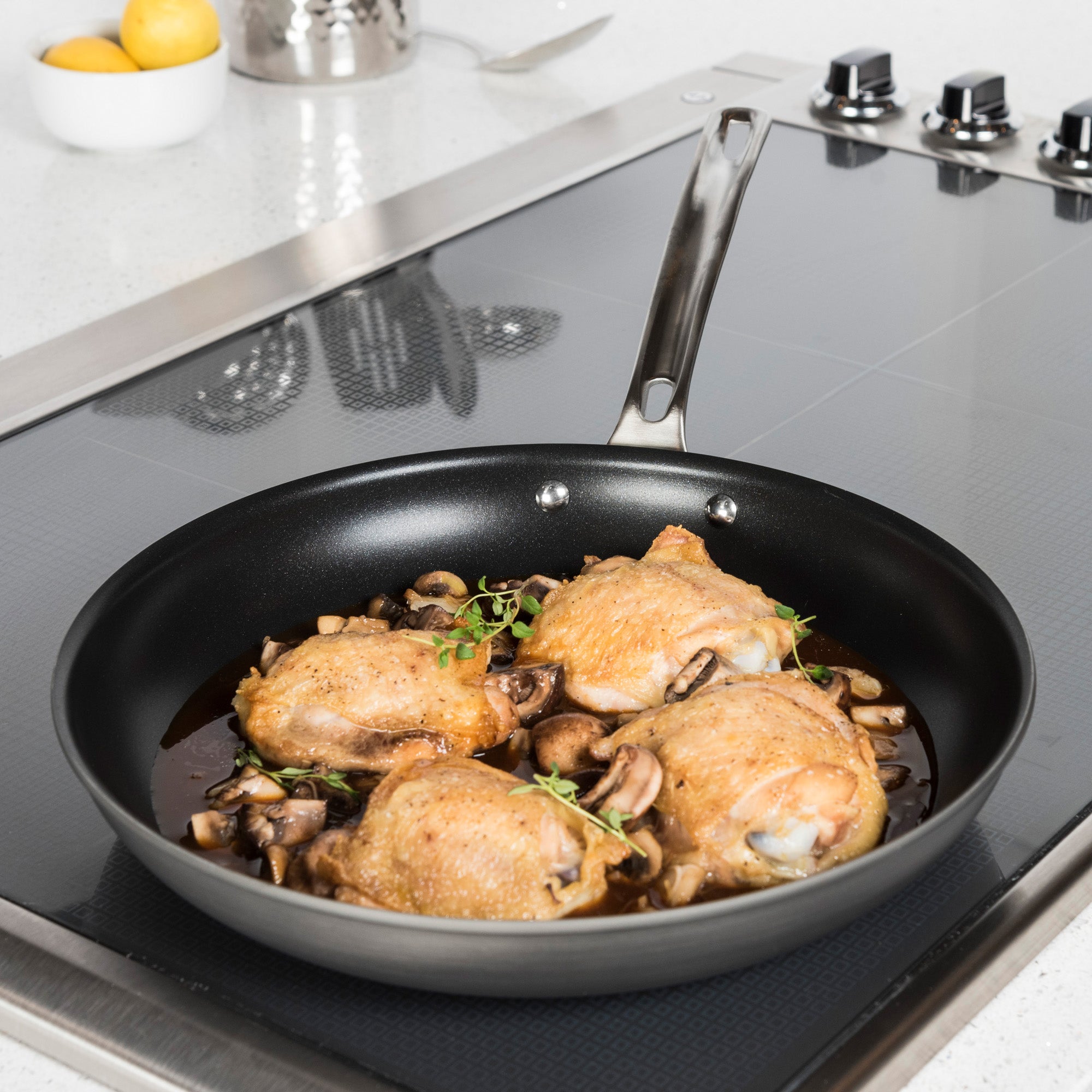 A Viking Hard Anodized Nonstick 12-Inch Fry Pan by Viking Culinary Products sits on the stove, filled with chicken thighs, mushrooms, and herbs in brown sauce. Its clean nonstick interior enables easy release; a bowl of lemons adds brightness in the background.