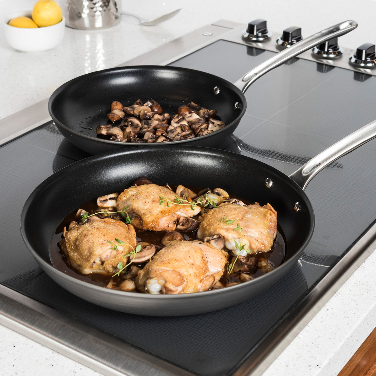 The Viking Hard Anodized Nonstick 2-Piece Fry Pan Set (10" & 12") by Viking Culinary Products is shown on a stovetop: one pan contains chicken thighs with mushrooms and herbs, the other sautéed mushrooms. Lemons and a pot are in the background.
