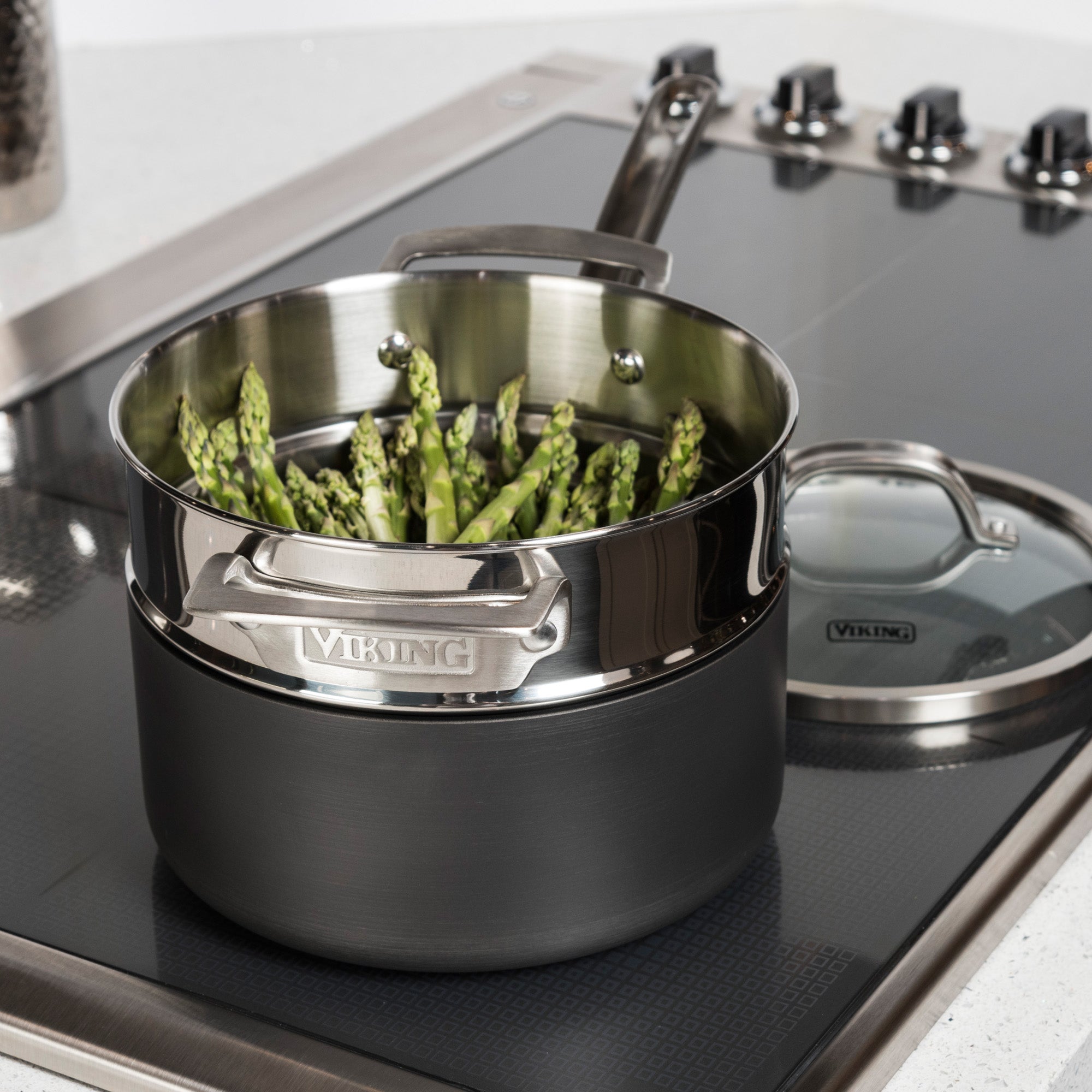 The Viking Stainless Steel Universal Steamer Insert and Colander by Viking Culinary Products rests beside a black hard anodized saucepan filled with upright asparagus on a sleek stovetop, its matching glass lid nearby.