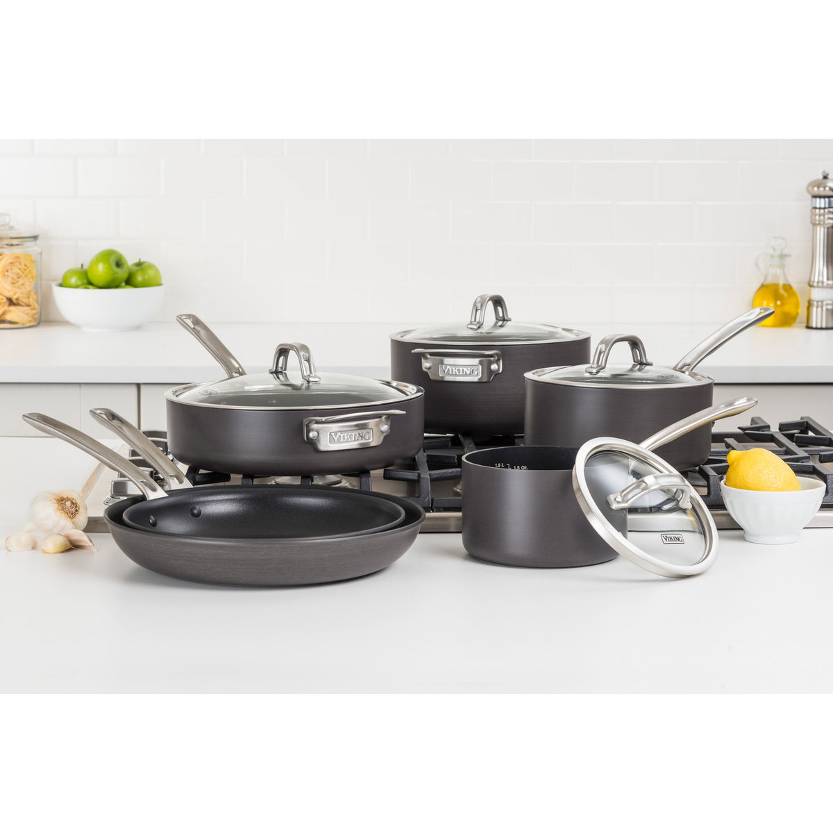 The Viking Culinary Products Hard Anodized Nonstick 10-Piece Cookware Set with glass lids is showcased on a white kitchen counter and stovetop, surrounded by garlic, lemons, apples, jars of pasta, and a Dutch oven.