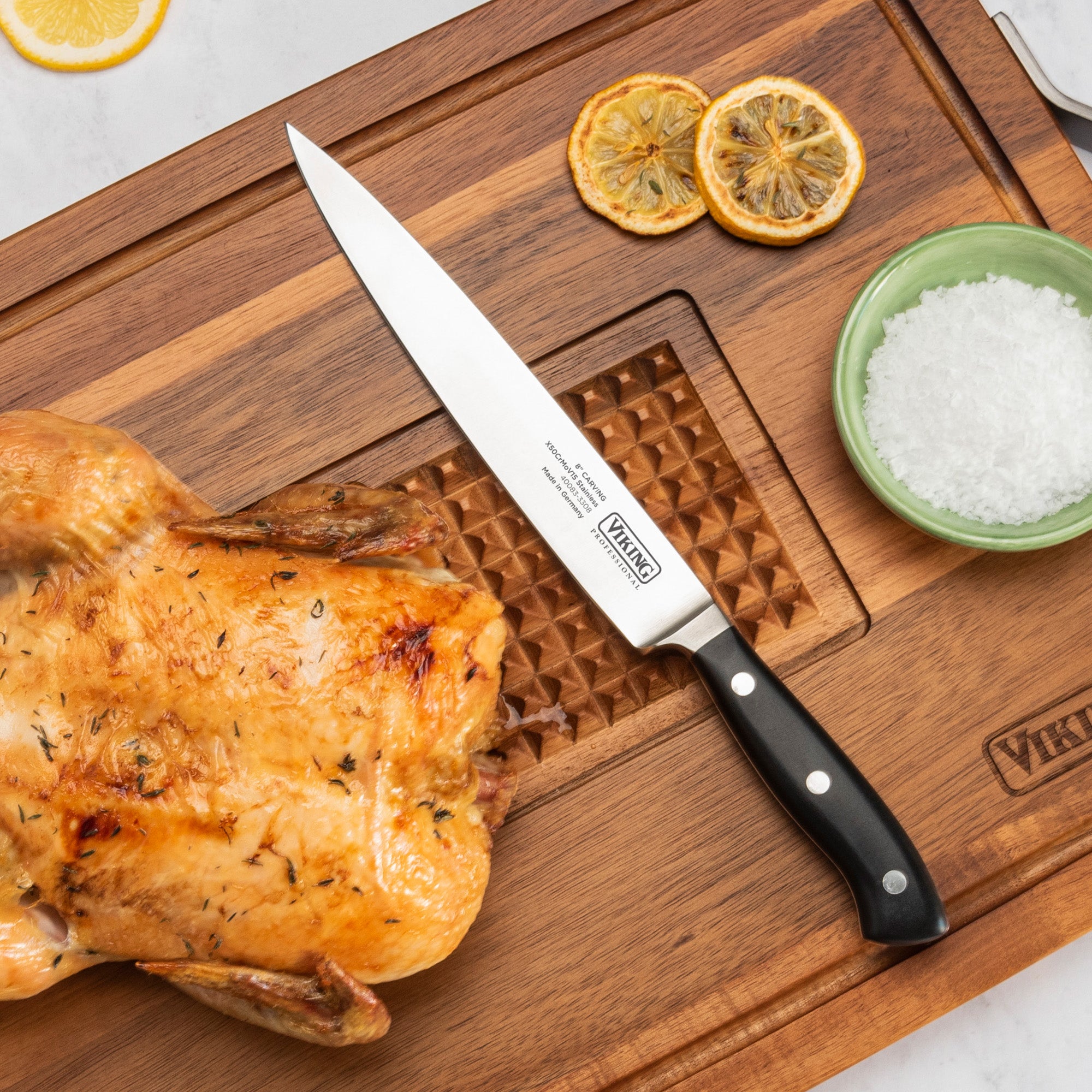 A roasted chicken rests on a wooden cutting board with a Viking Professional 8 Carving Knife by Viking Culinary Products, a green bowl of coarse salt, two dried lemon slices, and a groove for catching juices.