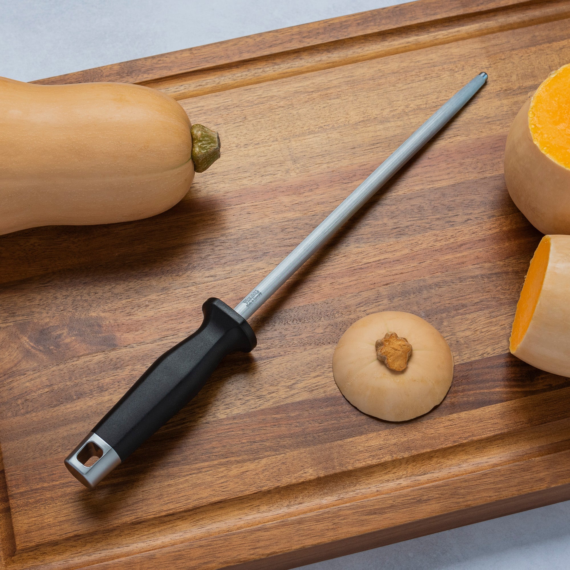 A Viking Professional 10-Inch Sharpening Steel by Viking Culinary Products with a black handle lies on a wooden cutting board beside a whole butternut squash, its top, and a halved butternut squash.
