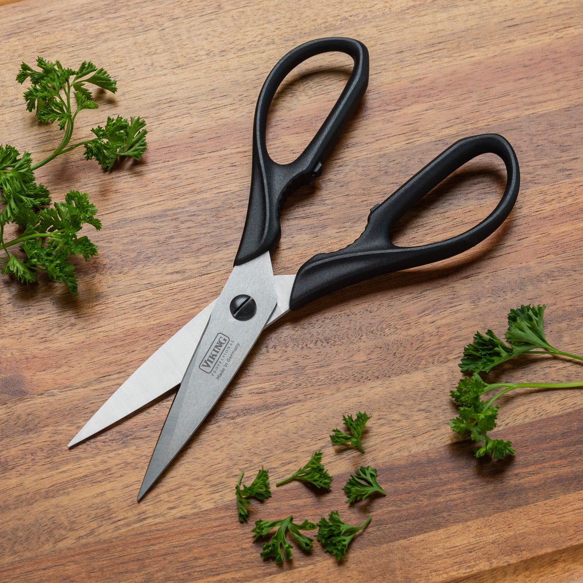 A pair of Viking Professional 8-Inch Scissors by Viking Culinary Products lies open on a wooden surface, surrounded by small sprigs of fresh parsley.