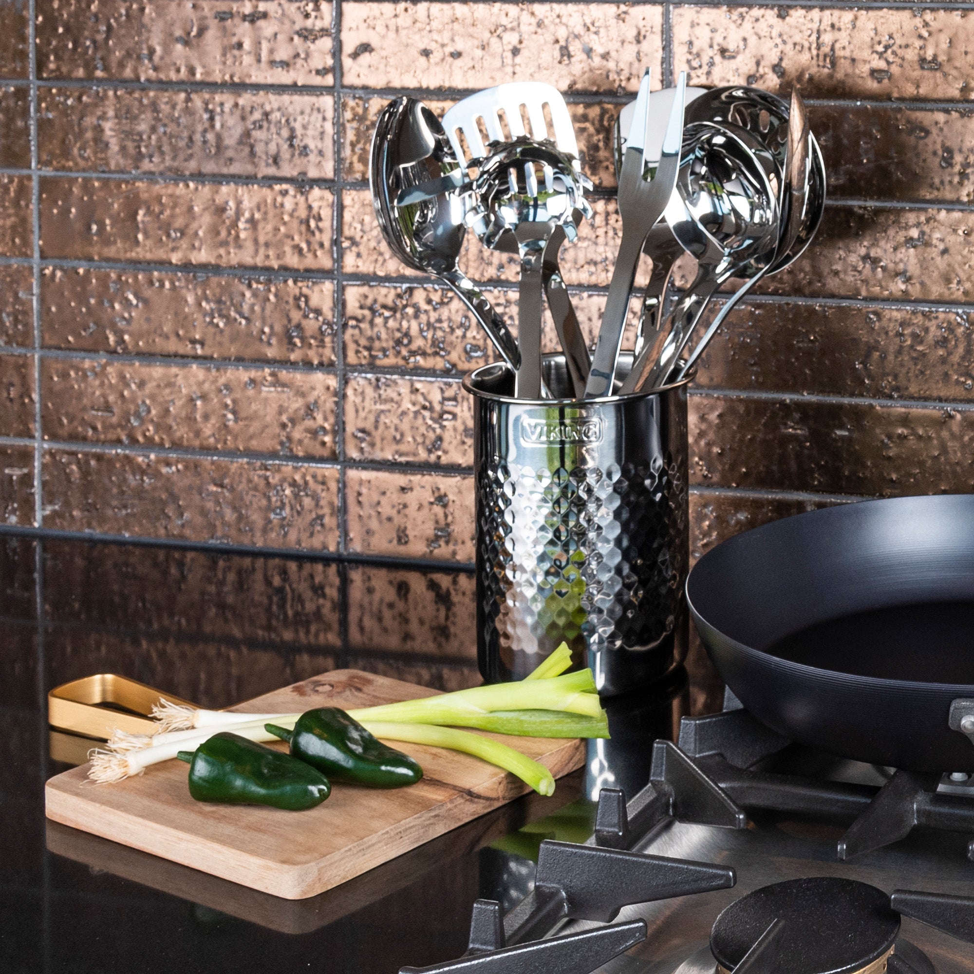 A Viking Hammered Stainless Steel Utensil Holder by Viking Culinary Products sits on a counter with kitchen tools beside a cutting board with green onions and poblano peppers, near a black skillet on the stove against brown tiled backsplash.