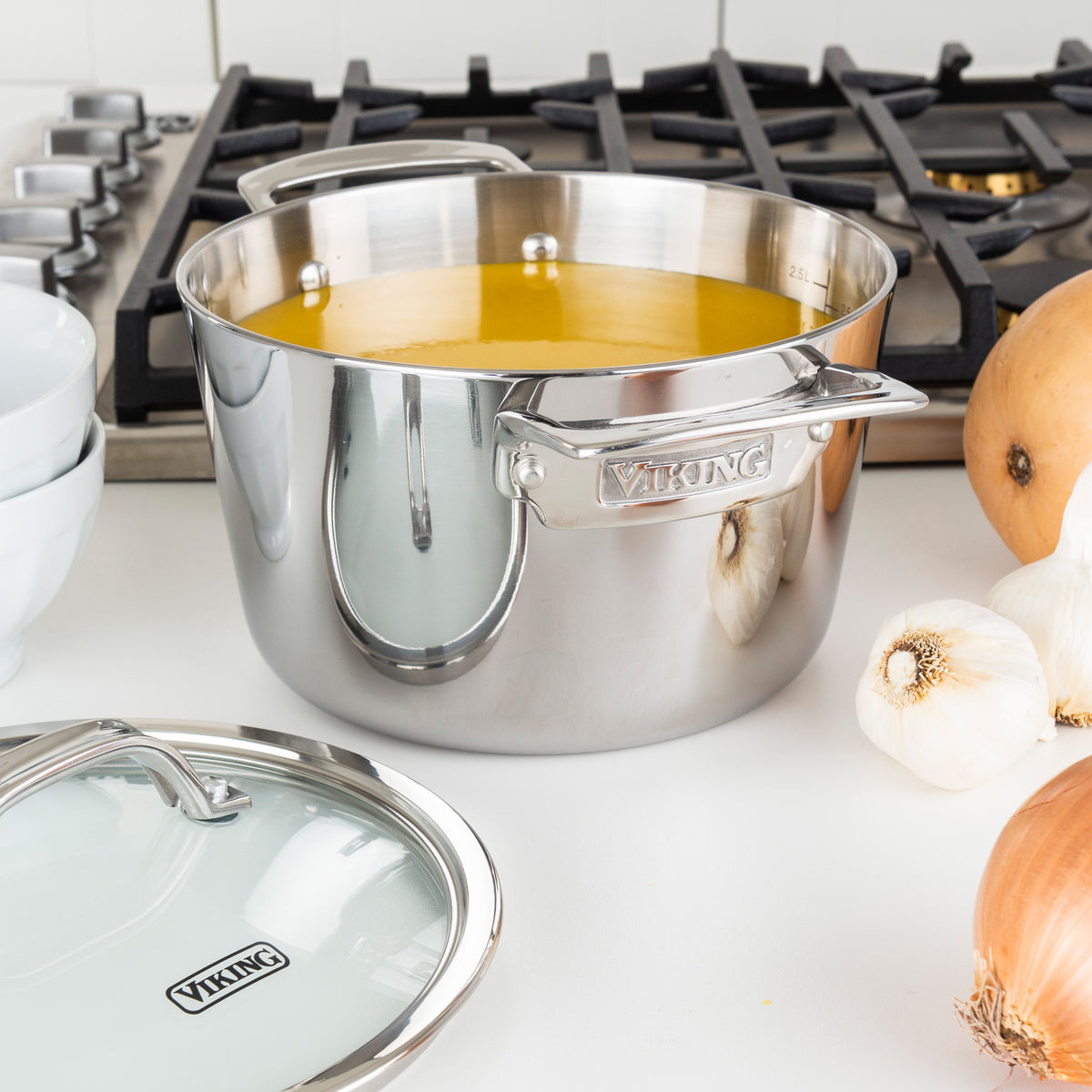 The Viking Contemporary 3-Ply Stainless Steel 3.4-Quart Soup Pot by Viking Culinary Products, filled with yellow soup, sits on a white countertop near a stove, surrounded by onions, garlic, and its glass lid.
