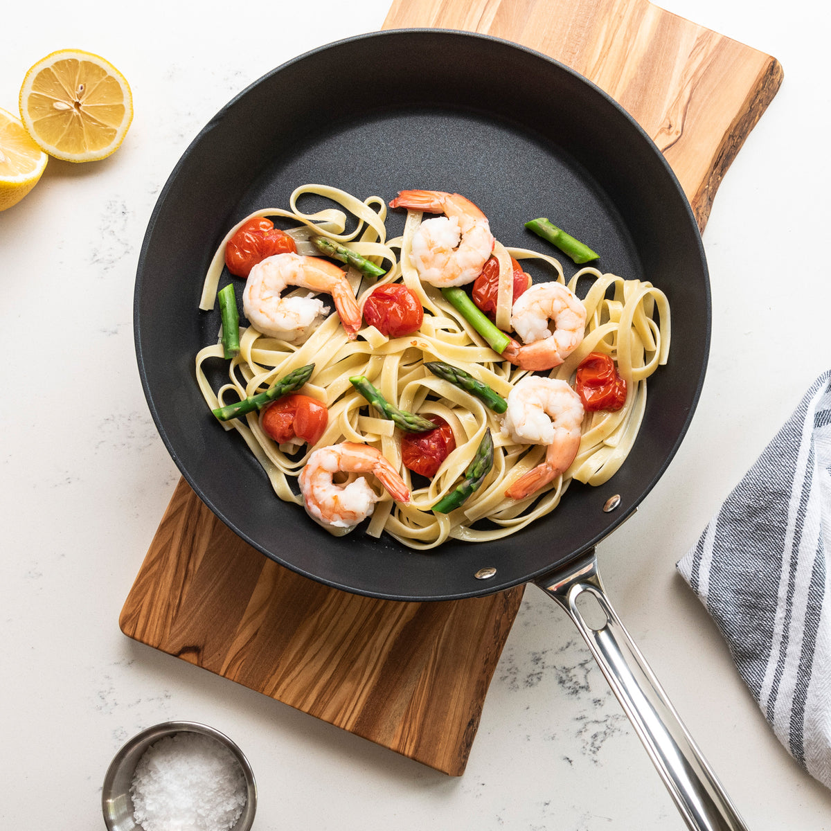 Shrimp pasta with cherry tomatoes and asparagus is served in a Viking Contemporary 3-Ply Stainless Steel 12-Inch Nonstick Fry Pan by Viking Culinary Products, alongside a halved lemon, salt bowl, and striped towel on a white counter.