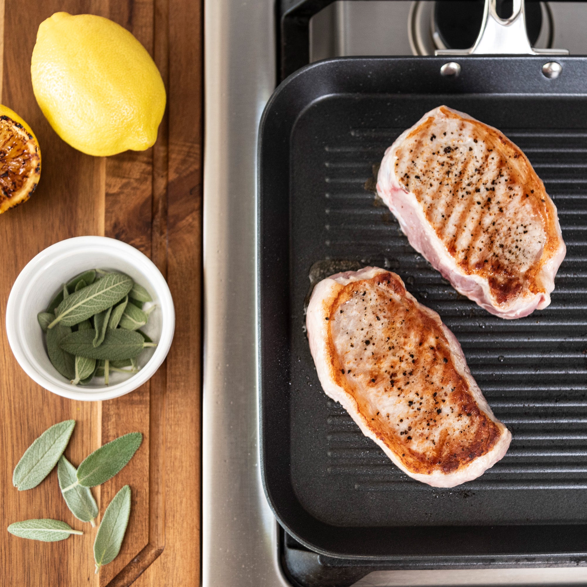 Two seasoned pork chops sizzle in the Viking Culinary Products Contemporary 3-Ply Stainless Steel 11-Inch Nonstick Grill Pan, while a lemon, grilled lemon half, and fresh sage leaves rest nearby—perfect for induction cooking.