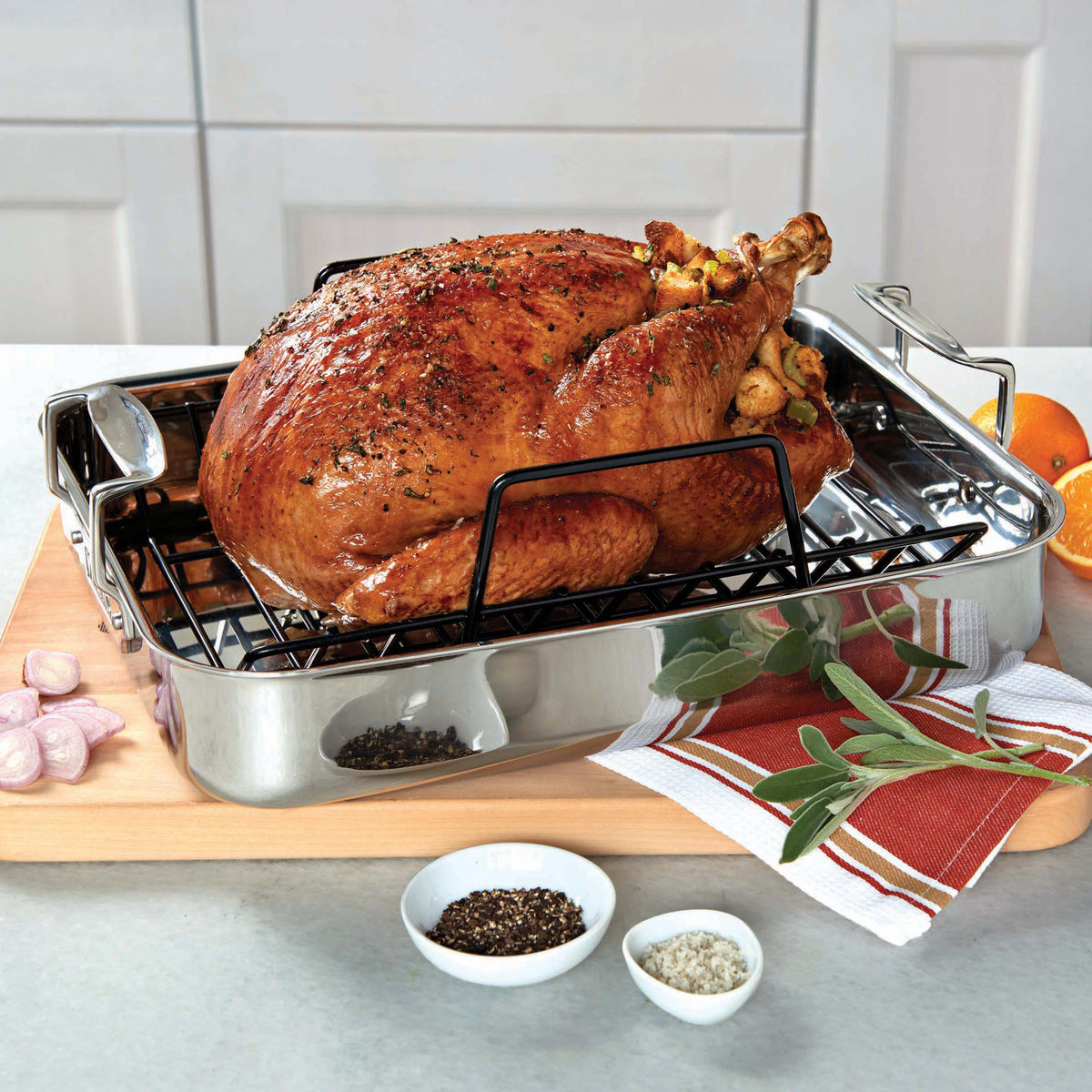 A roasted turkey rests on the Viking 3-Ply Stainless Steel Roaster with Rack by Viking Culinary Products, set on a wooden board and surrounded by fresh herbs, orange slices, shallots, and small bowls of salt and pepper.