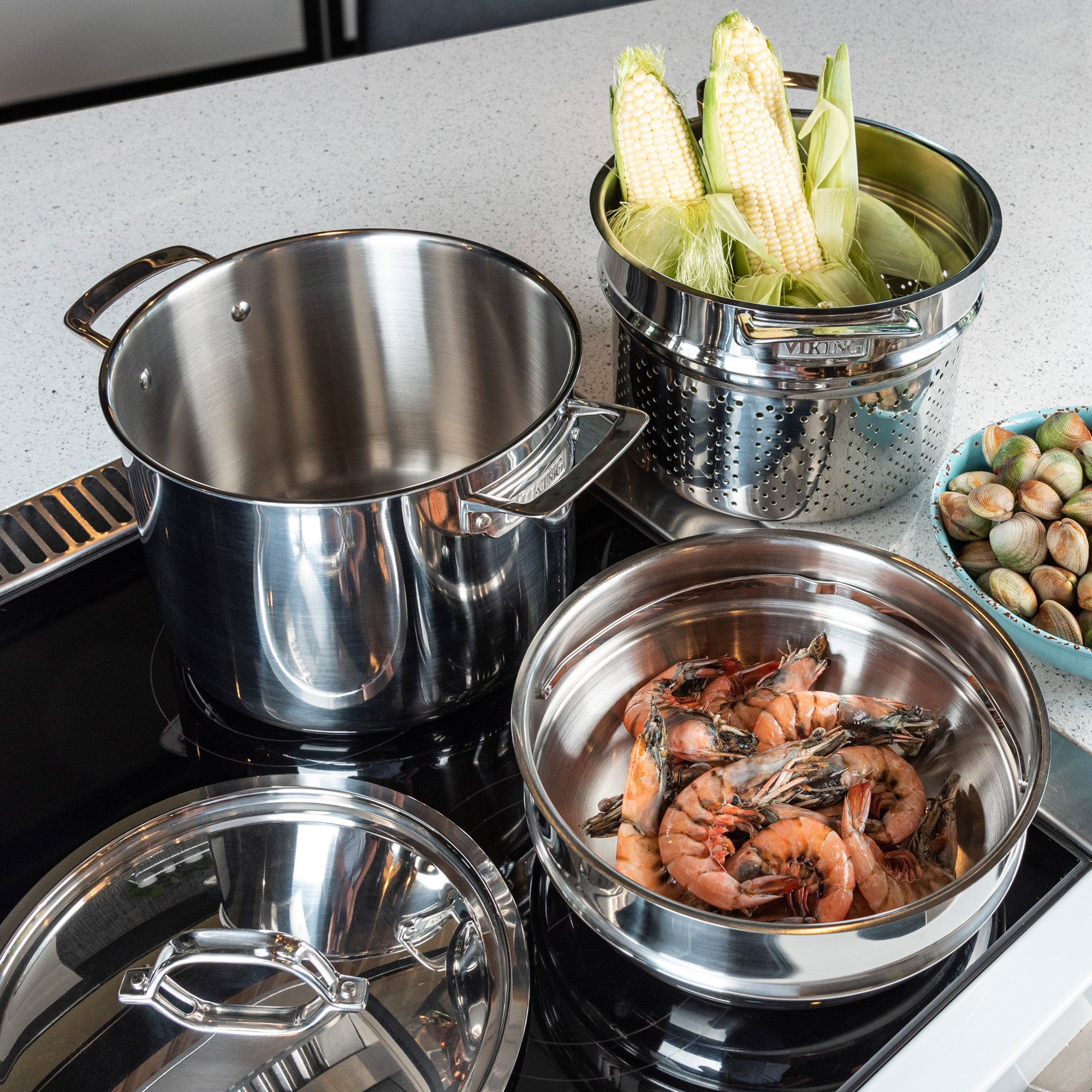 A kitchen scene with the Viking 3-Ply Stainless Steel 8 Qt Multipot 4-Piece Set by Viking Culinary Products on the stovetop; one pot holds shrimp in a pasta insert, another has corn, with clams nearby and both the metal lid and countertop visible.