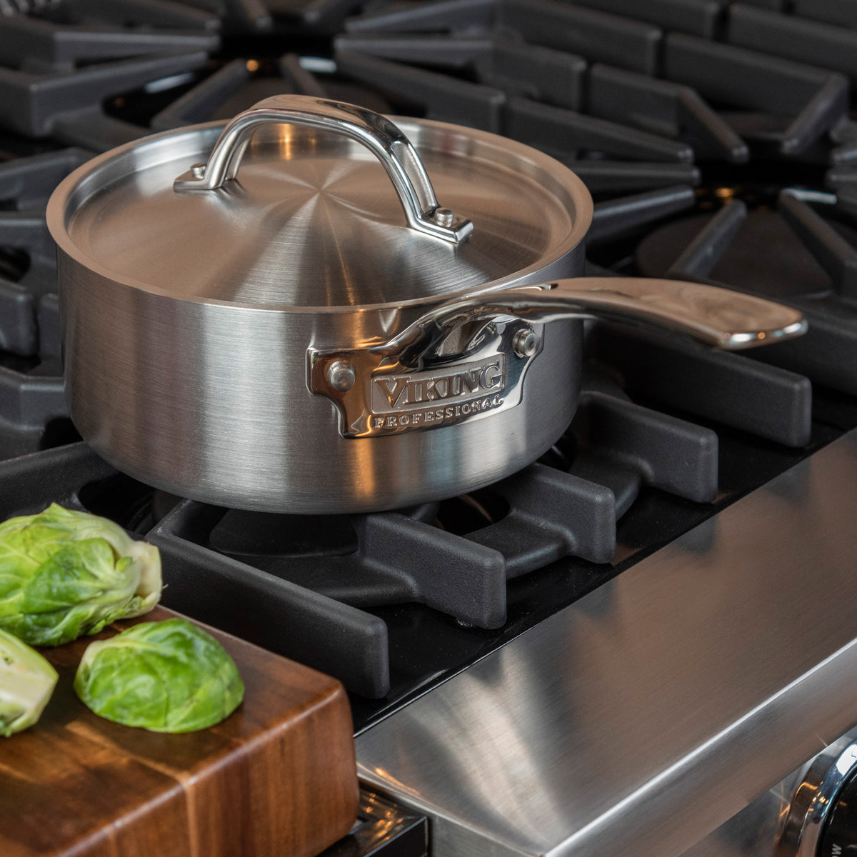 A Viking Professional 5-Ply 2-Quart Sauce Pan with Metal Lid by Viking Culinary Products sits on a gas stove beside a wooden cutting board topped with brussels sprouts.