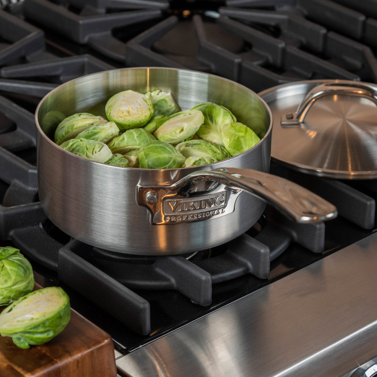 A Viking Professional 5-Ply 2-Quart Sauce Pan by Viking Culinary Products filled with halved Brussels sprouts sits on a gas stove, with its metal lid placed nearby.
