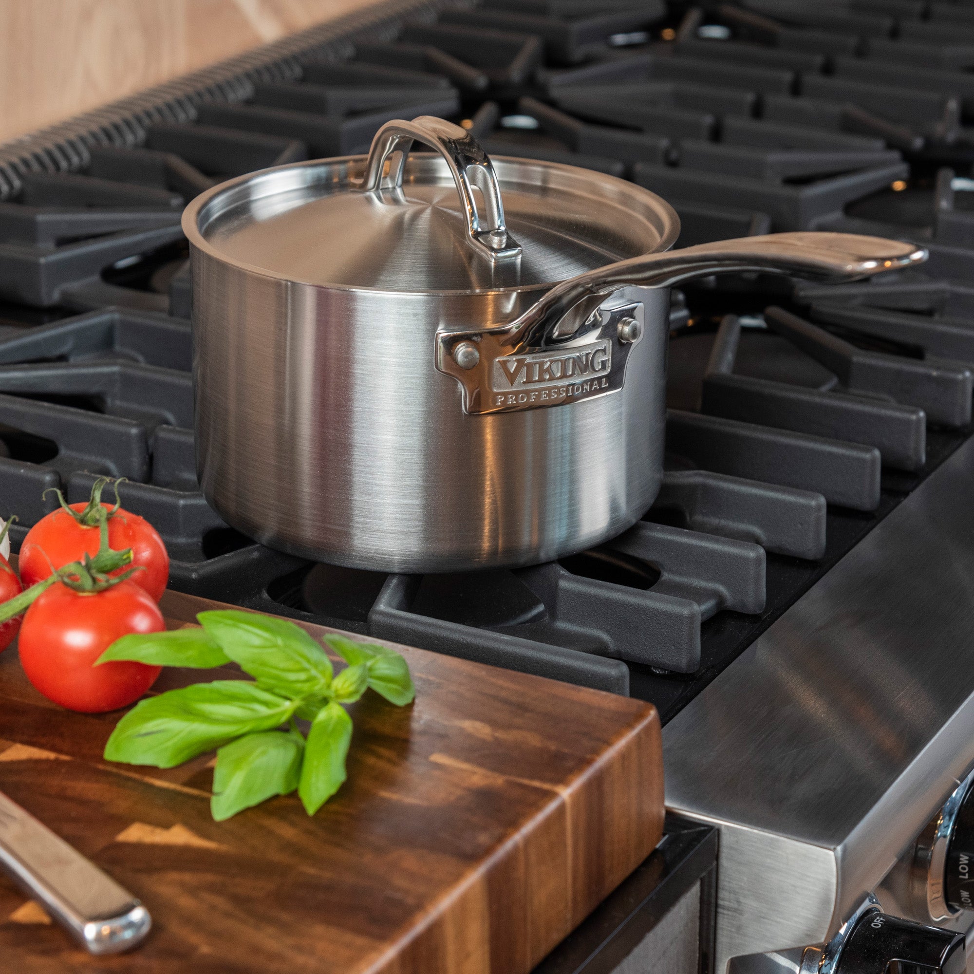 A Viking Culinary Products Professional 5-Ply 3-Quart Sauce Pan with a metal lid sits on a gas stove. Tomatoes and basil are on a wooden cutting board. This saucepan is dishwasher safe and ideal for making your favorite sauces.