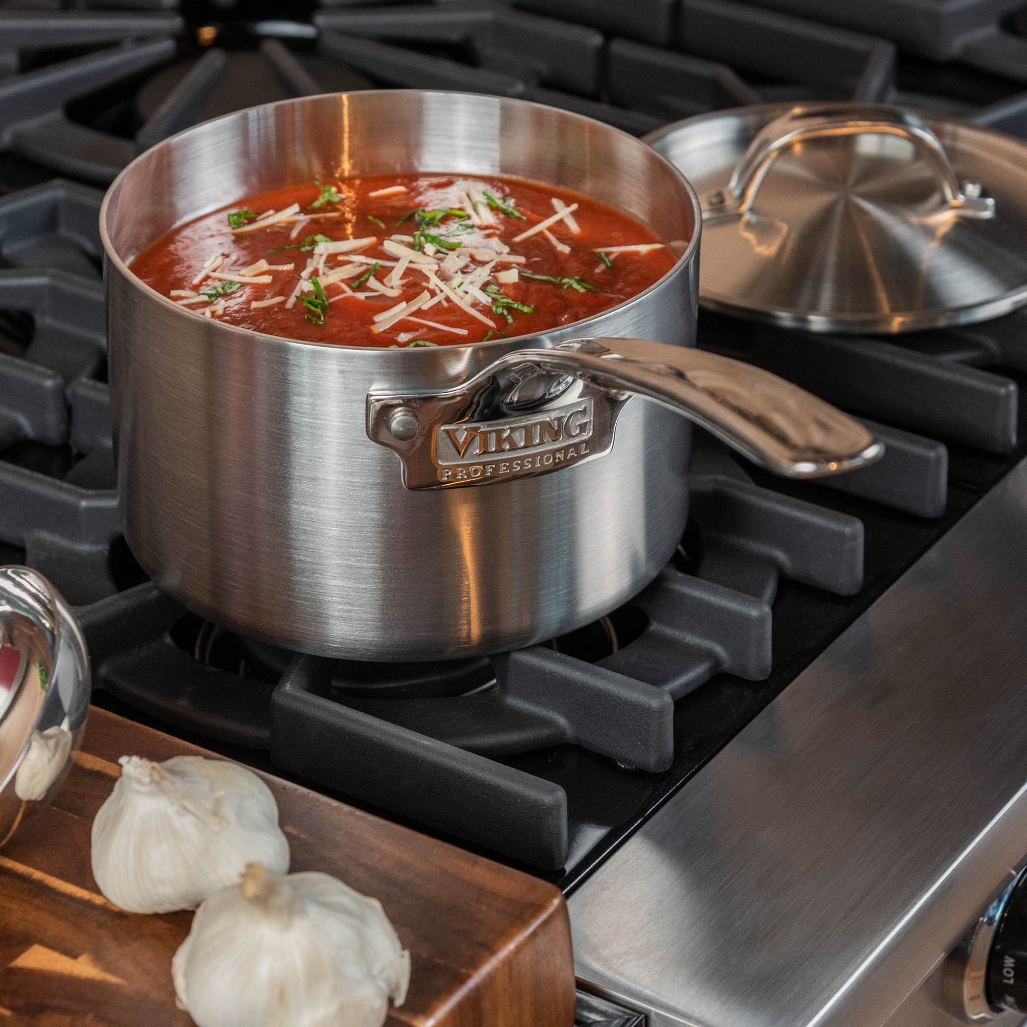The Viking Professional 5-Ply 3-Quart Sauce Pan with Metal Lid from Viking Culinary Products simmers tomato soup topped with herbs and cheese, while two garlic bulbs rest on a wooden board. Dishwasher safe for easy cleanup.