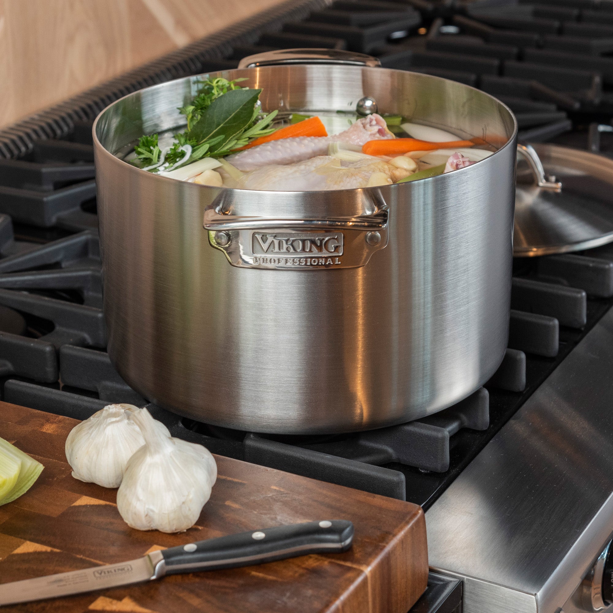 A Viking Culinary Products Viking Professional 5-Ply Stainless Steel 8-Quart Stock Pot filled with vegetables and chicken simmers on the stove, while fresh garlic bulbs and a knife rest on a wooden cutting board nearby. Induction compatible for even cooking.