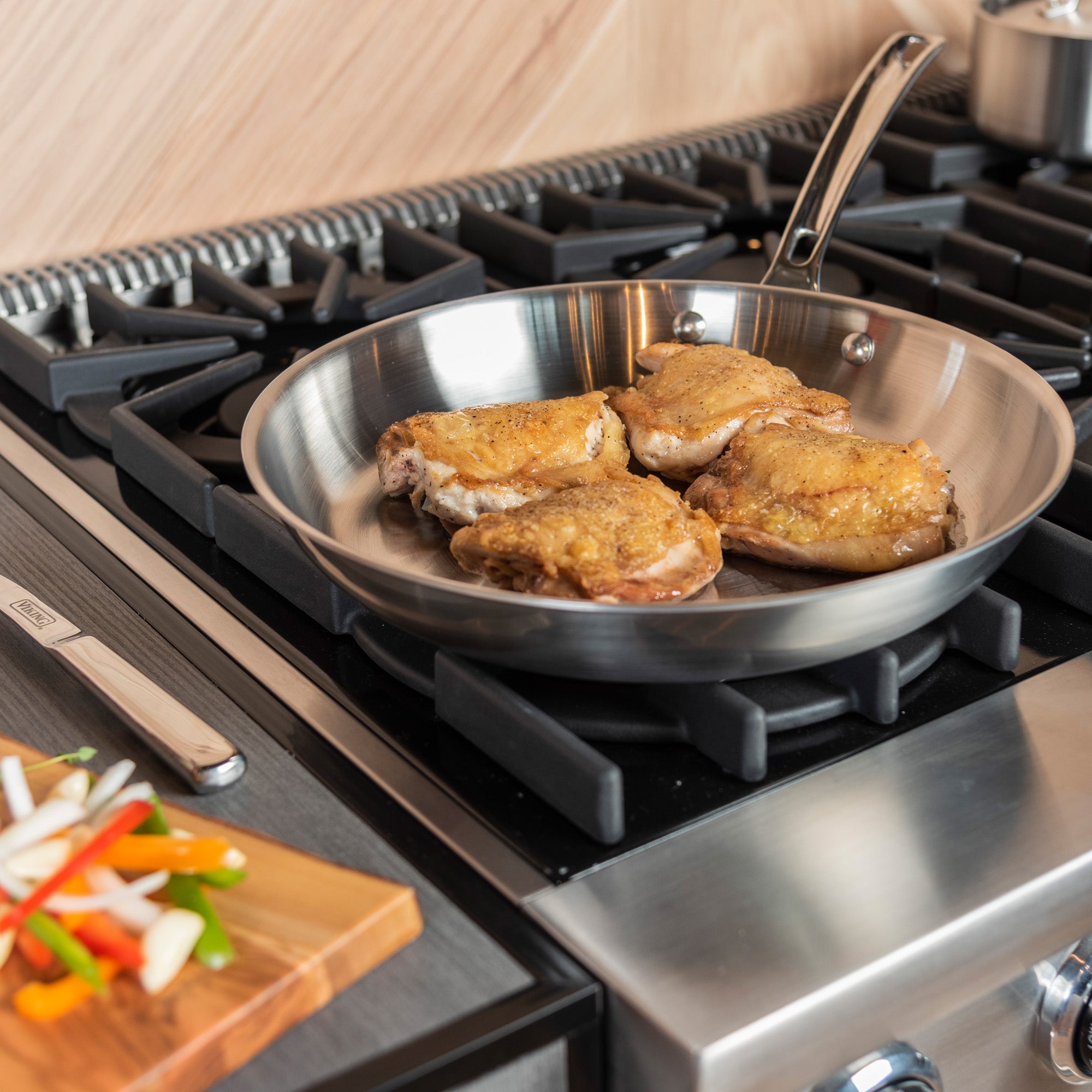 Chicken thighs cook in the Viking Professional 5-Ply 12-Inch Fry Pan from Viking Culinary Products on a gas stove. Sliced vegetables are on a wooden cutting board nearby. This stainless steel pan is dishwasher safe for easy cleanup.
