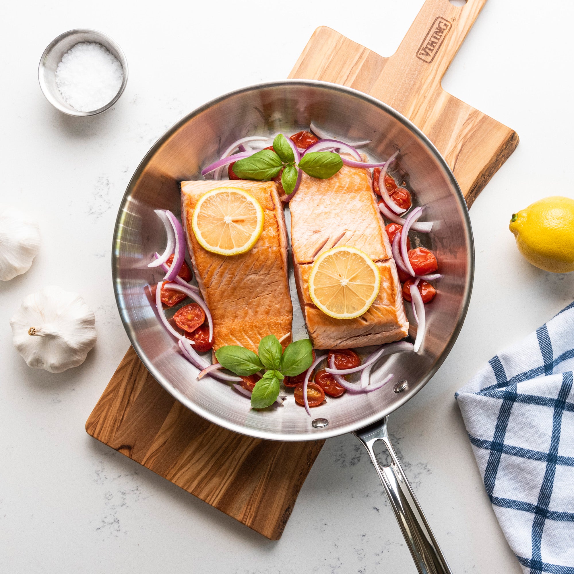 The Viking Professional 5-Ply 12-Inch Fry Pan by Viking Culinary Products holds two salmon fillets with lemon slices, cherry tomatoes, red onion, and basil. Next to it are garlic bulbs, a bowl of salt, a lemon, and a blue checkered towel on a wooden board.