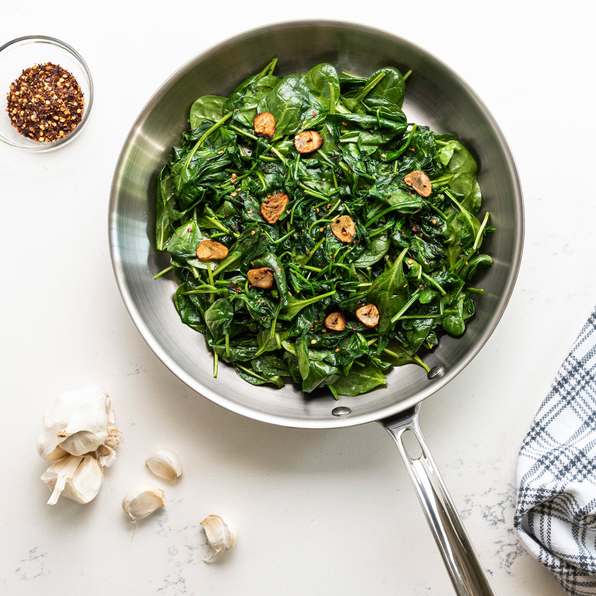 The Viking Professional 5-Ply 12-Inch Fry Pan by Viking Culinary Products, filled with spinach and garlic, sits on a white surface near red pepper flakes, garlic cloves, and a blue checkered towel. Dishwasher safe.