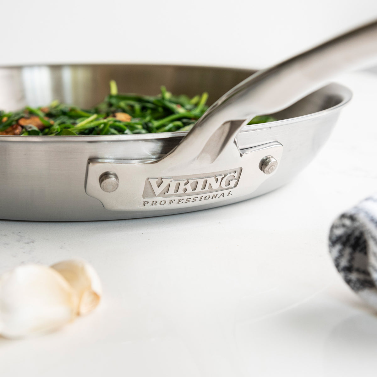 A close-up of the Viking Professional 5-Ply 12-Inch Fry Pan by Viking Culinary Products shows sautéed greens inside, with a garlic clove and a folded towel nearby. This stainless steel pan is also dishwasher safe for easy cleanup.