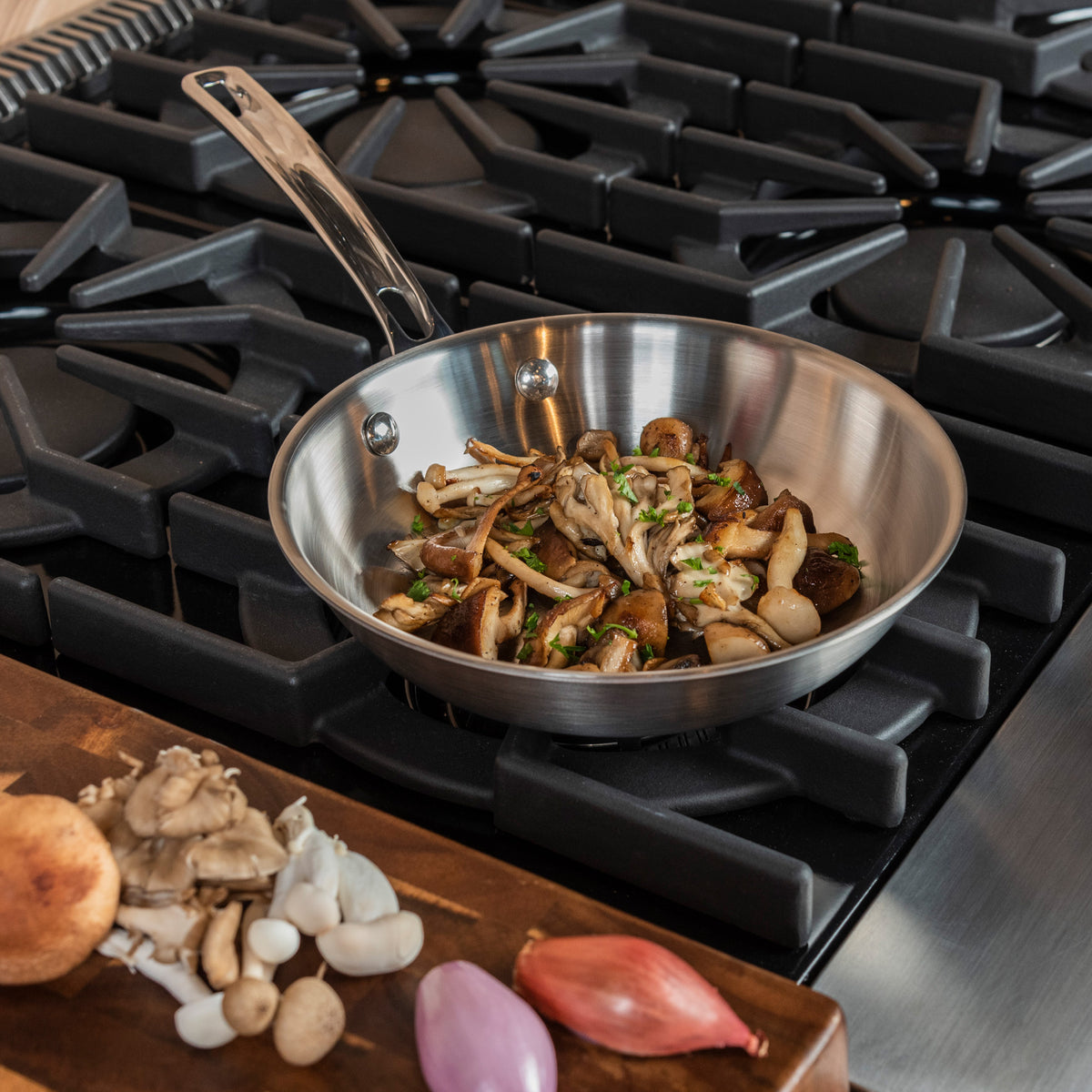 A Viking Professional 5-Ply 8-inch Fry Pan by Viking Culinary Products with sautéed mushrooms sits on a stovetop, while raw mushrooms and a shallot rest on a cutting board. This dishwasher safe pan simplifies meal prep.